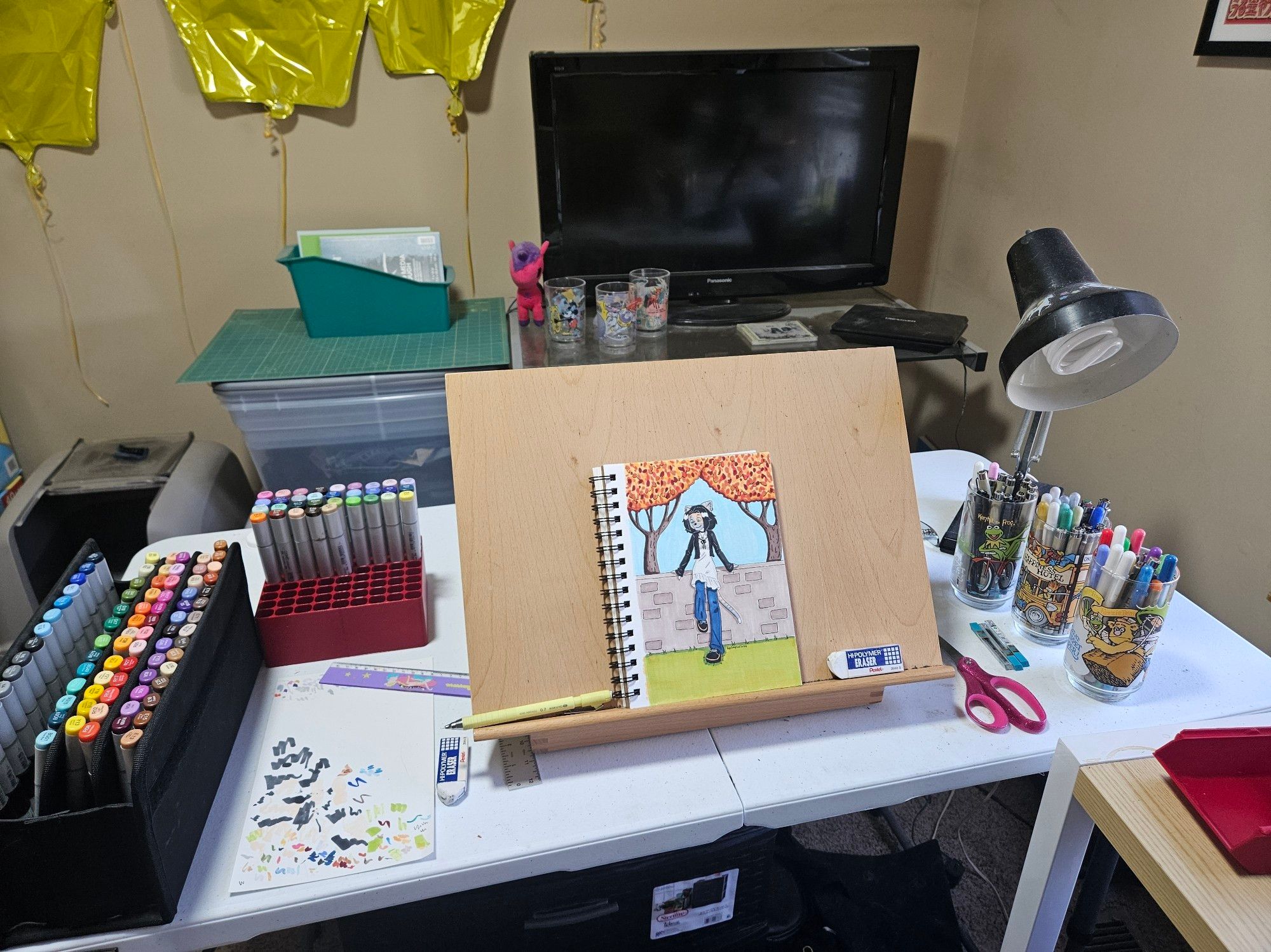 A folding table with a tabletop easel on it, with two storage containers worth of copics, some Muppet cups with pens in them, and a TV in the background