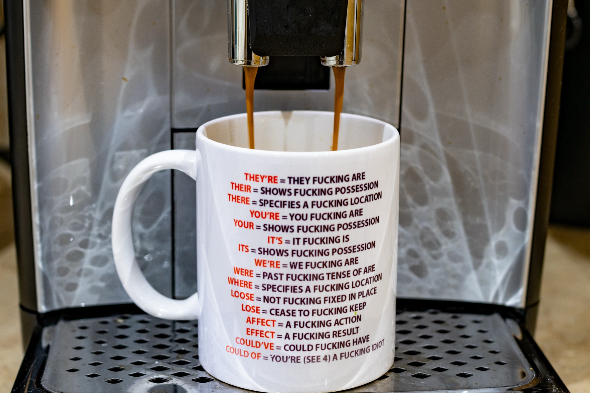 Coffee being poured into a mug from a machine. The mug is inscribed 

THEY'RE = THEY FUCKING ARE
THEIR = SHOWS FUCKING POSSESSION
THERE = SPECIFIES A FUCKING LOCATION
YOU'RE = YOU FUCKING ARE
YOUR = SHOWS FUCKING POSSESSION
IT'S = IT FUCKING IS
ITS = SHOWS FUCKING POSSESSION
WE'RE = WE FUCKING ARE
WERE = PAST FUCKING TENSE OF ARE
WHERE = SPECIFIES A FUCKING LOCATION
LOOSE = NOT FUCKING FIXED IN PLACE
LOSE = CEASE TO FUCKING KEEP
AFFECT = A FUCKING ACTION
EFFECT = A FUCKING RESULT
COULD OF = YOU’RE (SEE 4) A FUCKING IDIOT