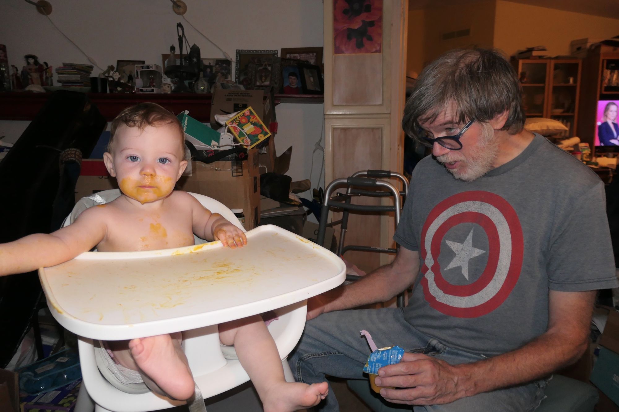 A seething nine-month old blue-eyed child with gooey yellow stuff all over her face, being fed by Mr. Space in a Captain America shirt.