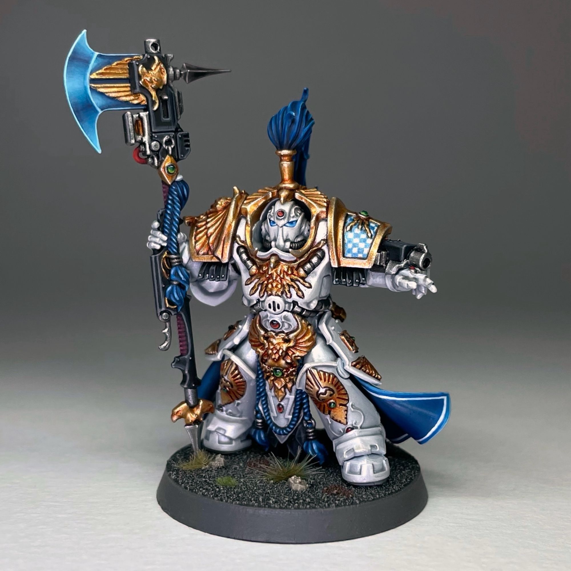 An Adeptus Custodes Allarus terminator for Warhammer 40,000 painted in a metallic white and gold armour with blue robes and accessories. The blade of the axe is painted with a blue non metallic metal effect. The right shoulder pad is a gold eagle while the left one has metallic blue and white checks.