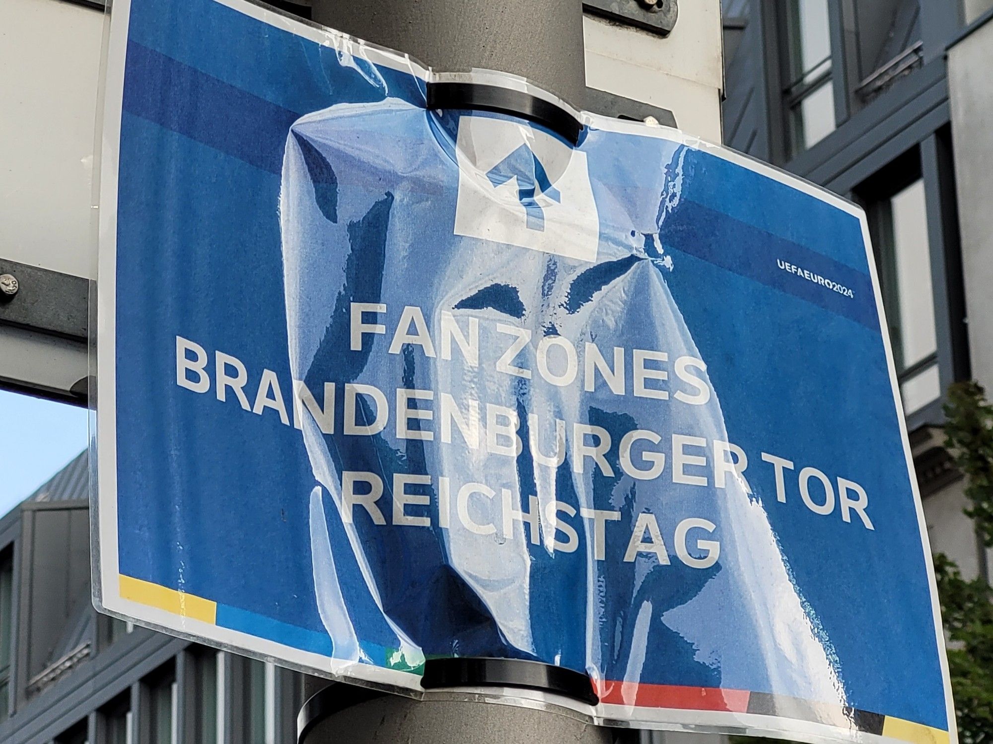 Blaues Schild an einem Laternenpfahl mit einem Pfeil und der Aufschrift "Fanzones Brandenburger Tor / Reichstag". Sehr klein im Eck das Logo der Fußball-Europameisterschaft 2024.