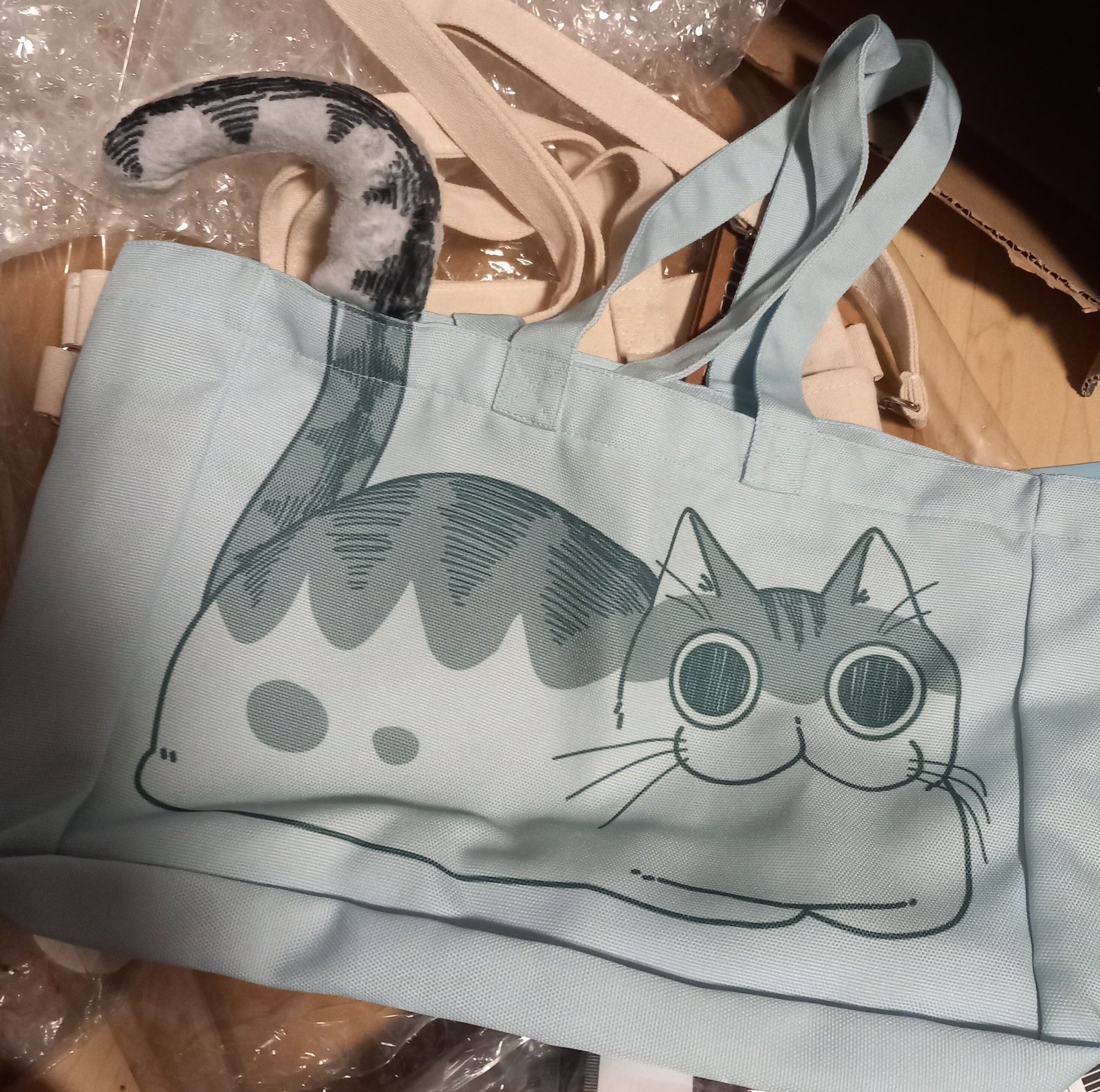 A light blue handbag lays in the center of the picture.
On the bag is print of a white cat with a gray back and black stripes.
The cat looks directly at you and their mouth looks like a turned three. Their eyes a completely round like a circle and their pupils are big and nearly fill out the eyes completely.

The cats is printed in a lying down pose and their paws are safely tucked under their body. The tail breaks the limited space on the front of the back and a extended tail got added on the rim of the back. Because of this it's looks like the tail continues outside the back.