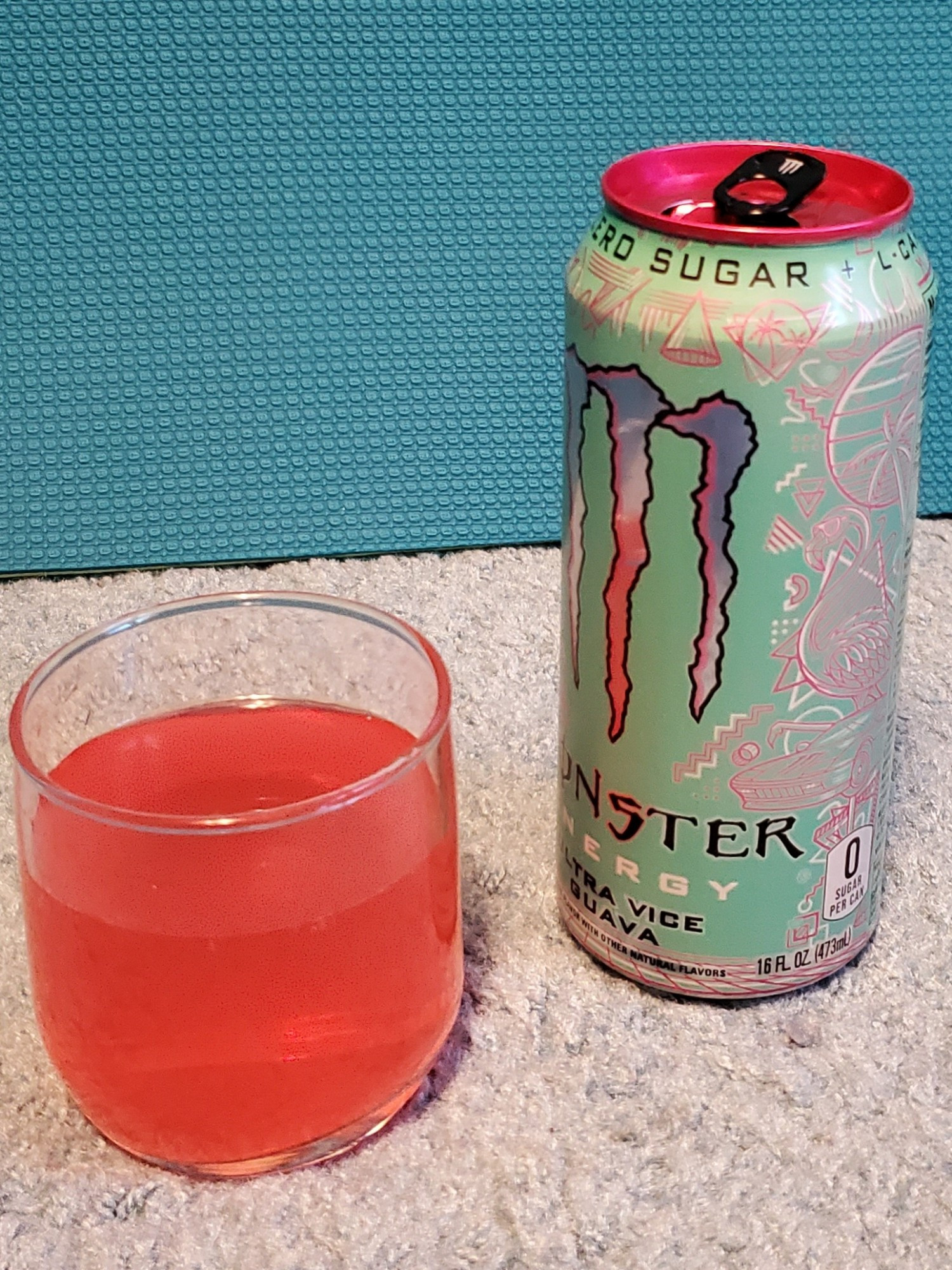 A short glass with hot pink liquid next to a Monster Energy can for ultra vice guava flavor. The can has a light turquoise or seafoam green background, silver lettering for the M monster logo, and various pink outlines of retro beach city scenes, an '80s car, flamingos, and geometric shapes. The background is a light blue carpet, with the objects set in front of a teal backboard.