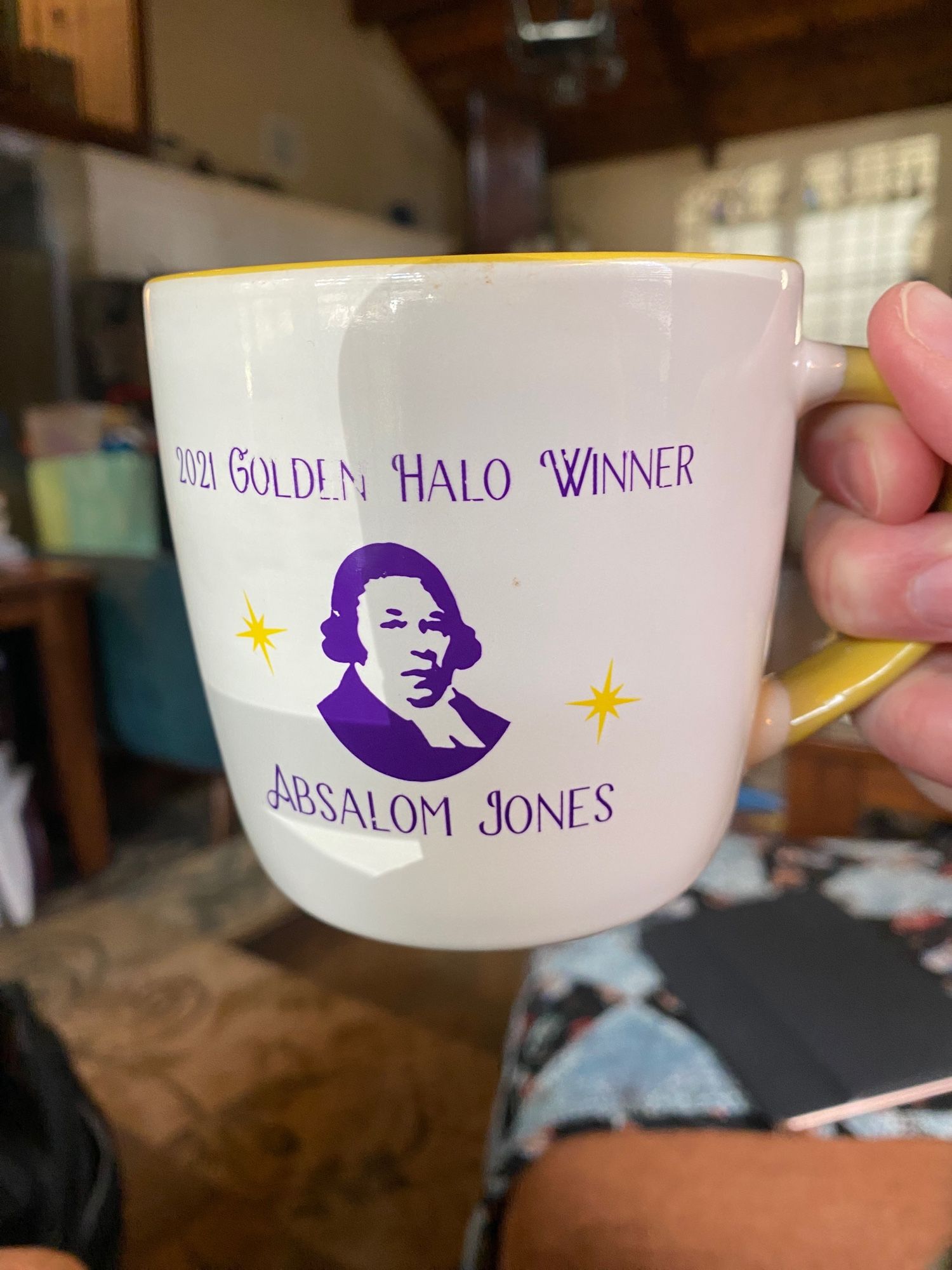 Hand (mine) holding up a white mug with yellow handle and accents with purple print reading “2021 Golden Halo Winner Absalom Jones” above and below a purple printed rendering of Philadelphia abolitionist Absalom Jones.