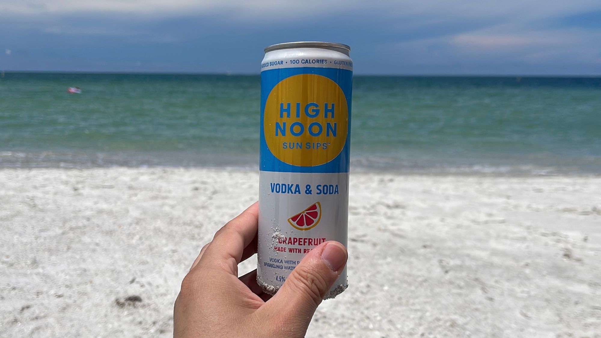 A can of grapefruit High Noon held in front of the beach meeting the ocean. The horizon lines up with top of the blue label while the shoreline lines up with the bottom of the label.