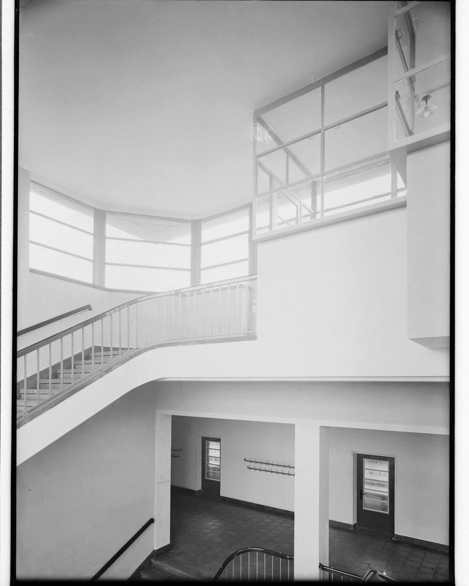 Ein Innenblick auf ein modern anmutendes Gebäude aus dem Jahr 1930 mit einer geschwungenen Treppe, großen Fenstern und minimalistischen Designelementen. Das Schwarz-Weiß-Bild zeigt klare Linien und offene Räume und hebt den architektonischen Stil hervor.