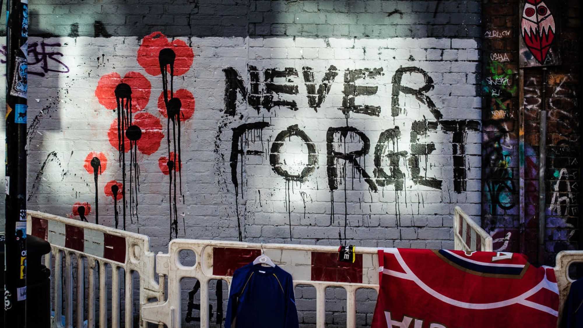 Eine Backsteinmauer zeigt Graffiti mit dem Text "NIE VERGESSEN" neben roten Mohnblumen mit tropfender Farbe. Eine Baubarriere und Kleidung hängen vor der Wand.