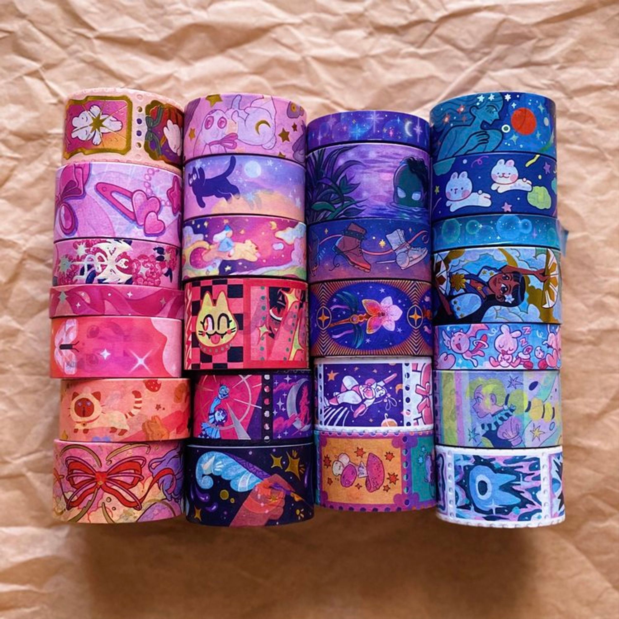 Photo showing the entire Magical Girl washi tape collection, consisting of 26 designs created by 24 artists from around the world. The tapes are organized by color and come in various styles and finishes. The color palette of the collection is predominantly pink, purple, and blue.