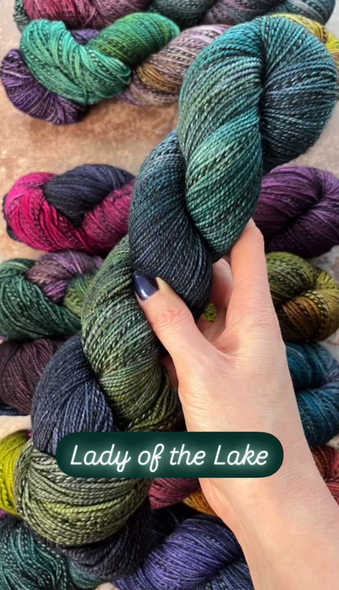 A white woman’s hand with dark purpleish-blue painted nails hold a skein of hand dyed sock yarn. The yarn has a black and grey zebra style marl running through it, and is dyed in shades of deep navy blue, teal and moss green. A caption on the photo says the yarn shade is called Lady of the Lake.