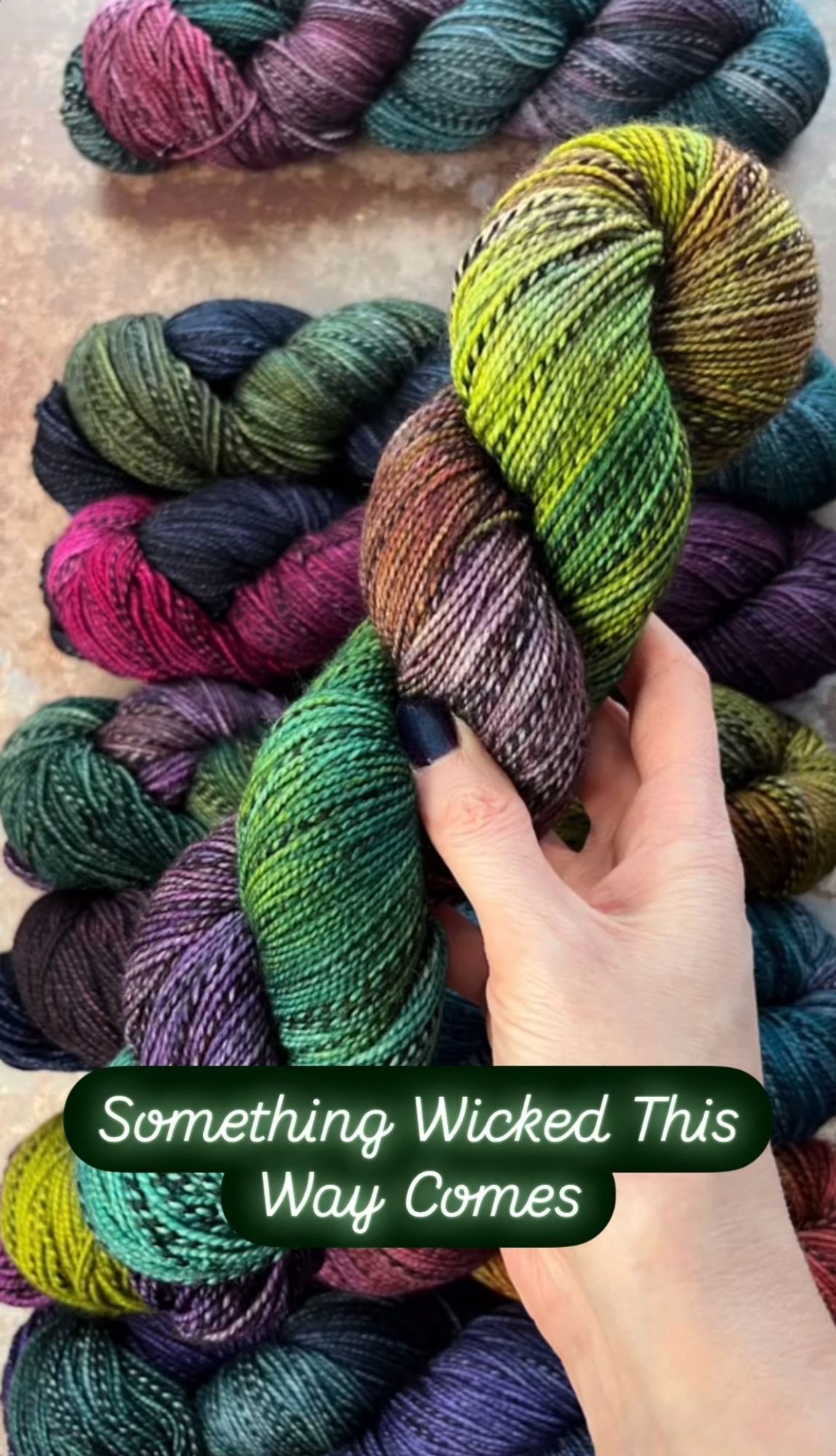 A white woman’s hand with dark purpleish-blue painted nails hold a skein of hand dyed sock yarn. The yarn has a black and grey zebra style marl running through it, and is dyed in shades of jade green, yellow and purple. A caption on the photo says the yarn shade is called Something Wicked This Way Comes.