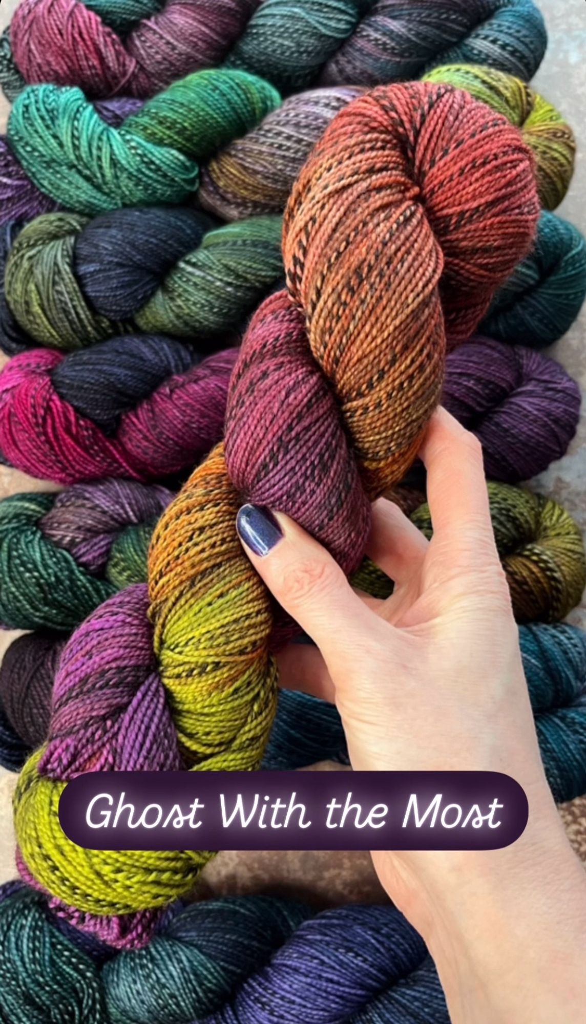 A white woman’s hand with dark purpleish-blue painted nails hold a skein of hand dyed sock yarn. The yarn has a black and grey zebra style marl running through it, and is dyed in shades of vivid lime green, rusty orange and bright warm purple. A caption on the photo says the yarn shade is called Ghost With the Most.