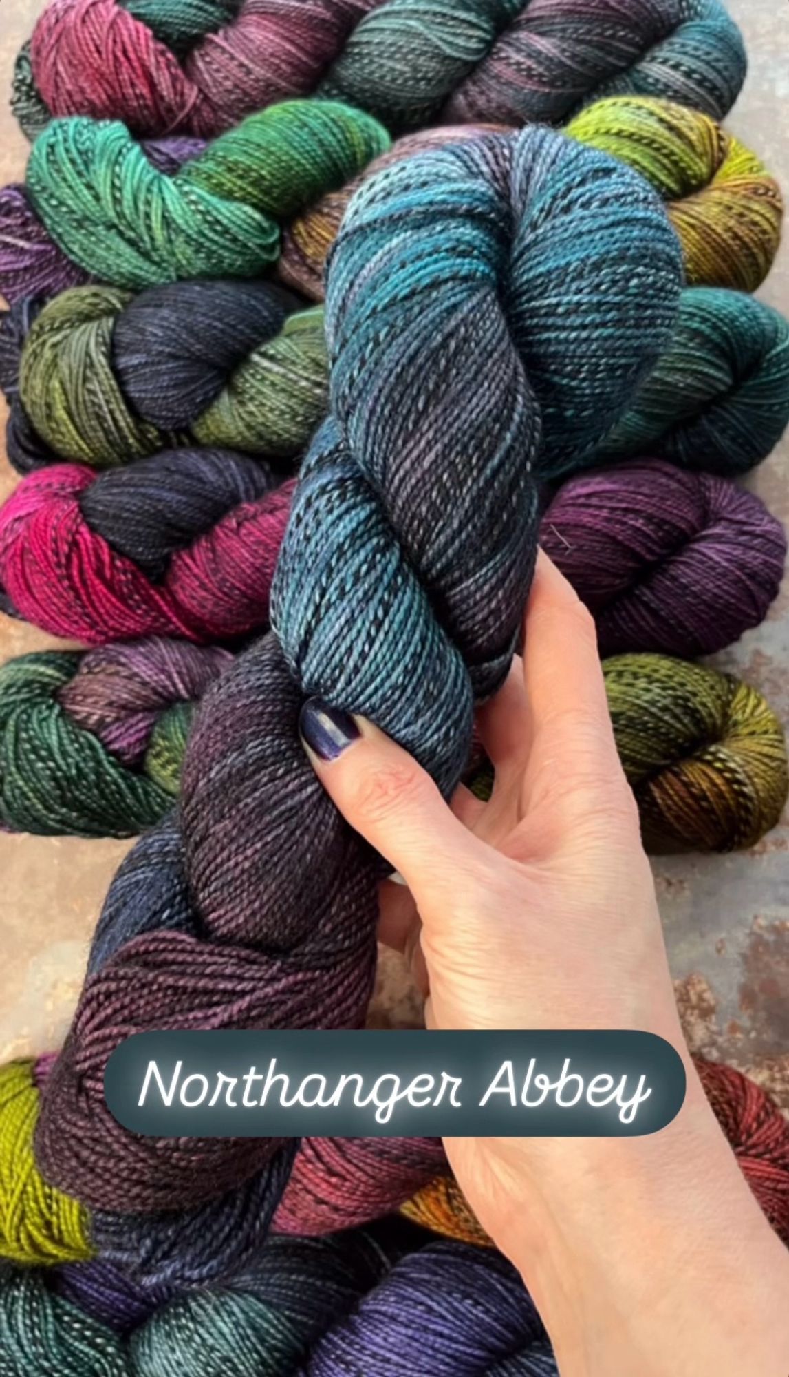 A white woman’s hand with dark purpleish-blue painted nails hold a skein of hand dyed sock yarn. The yarn has a black and grey zebra style marl running through it, and is dyed in shades of deep plum, smoky teal and navy blue. A caption on the photo says the yarn shade is called Northanger Abbey.