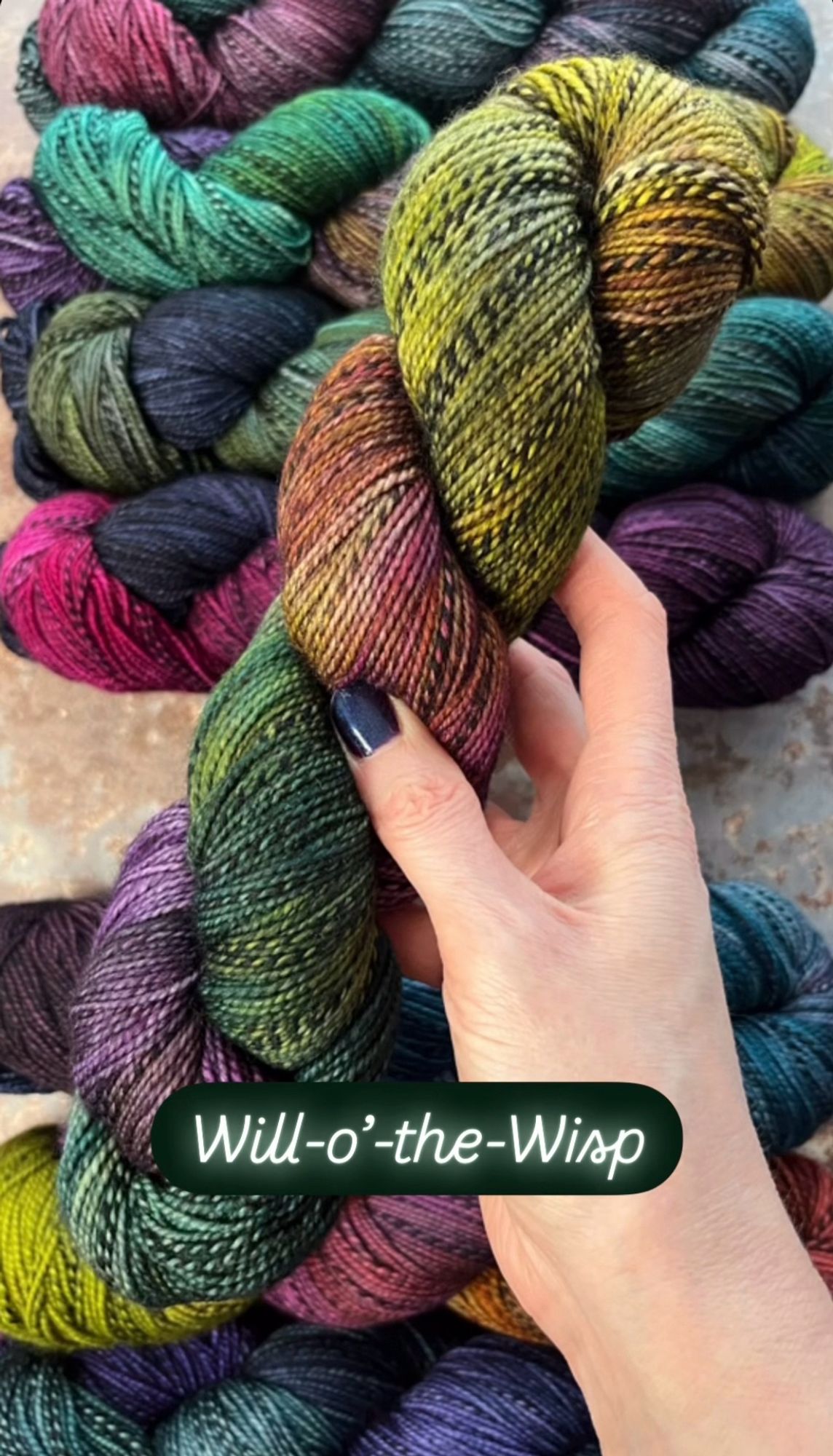 A white woman’s hand with dark purpleish-blue painted nails hold a skein of hand dyed sock yarn. The yarn has a black and grey zebra style marl running through it, and is dyed in shades of deep forest green, gold, rust and deep purple. A caption on the photo says the yarn shade is called Will-o’-the-Wisp.