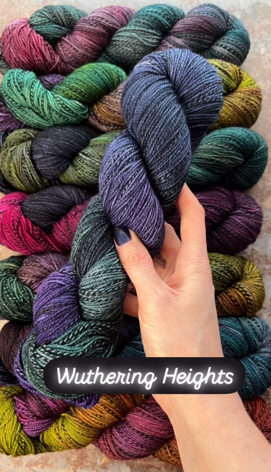 A white woman’s hand with dark purpleish-blue painted nails hold a skein of hand dyed sock yarn. The yarn has a black and grey zebra style marl running through it, and is dyed in shades of deep forest green, dark navy blue and deep violet. A caption on the photo says the yarn shade is called Wuthering Heights.