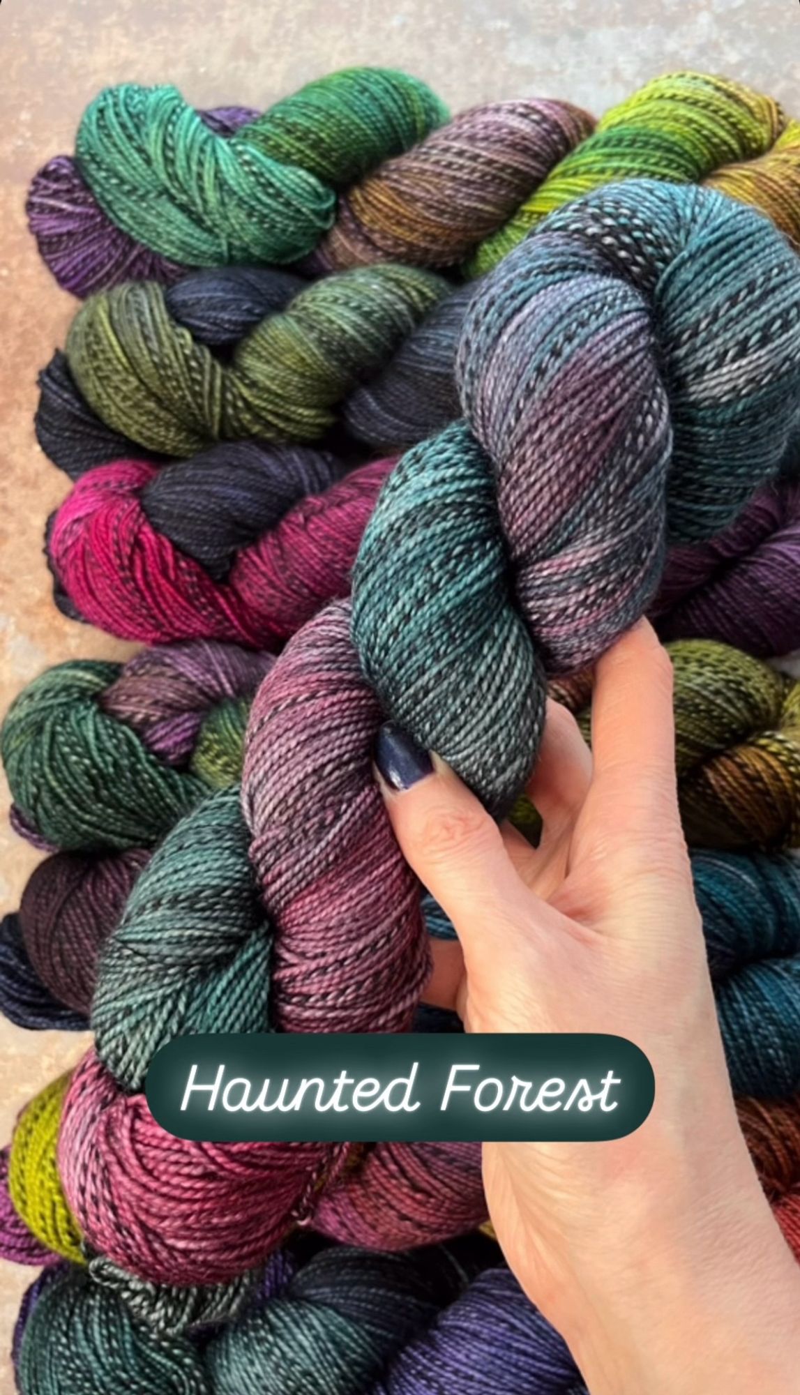 A white woman’s hand with dark purpleish-blue painted nails hold a skein of hand dyed sock yarn. The yarn has a black and grey zebra style mark running through it, and is dyed in shades of deep red, teal and forest green. A caption on the photo says the yarn shade is called Haunted Forest.