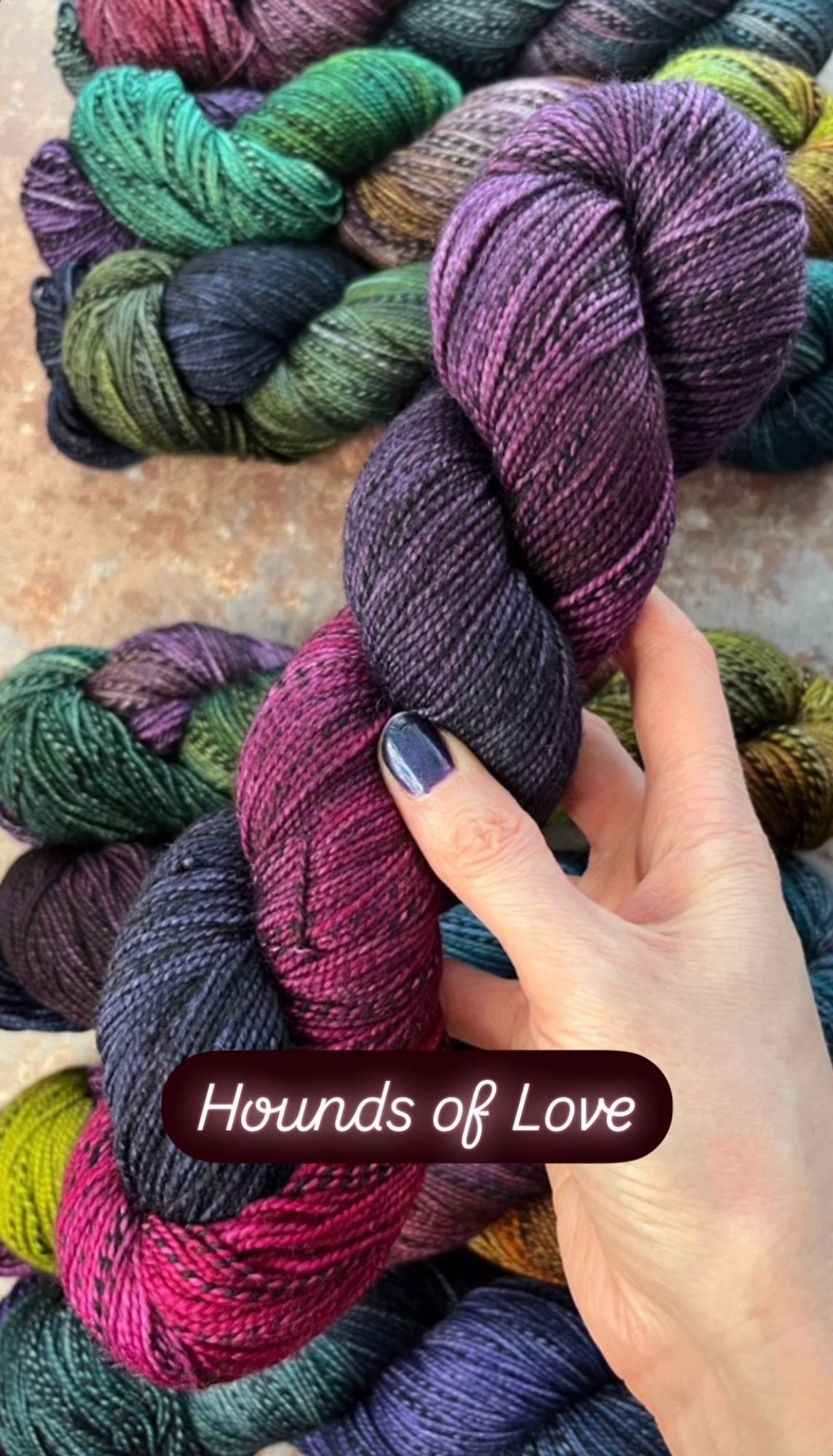 A white woman’s hand with dark purpleish-blue painted nails hold a skein of hand dyed sock yarn. The yarn has a black and grey zebra style mark running through it, and is dyed in shades of cerise pink, deep plum and dark navy. A caption on the photo says the yarn shade is called Hounds of Love.