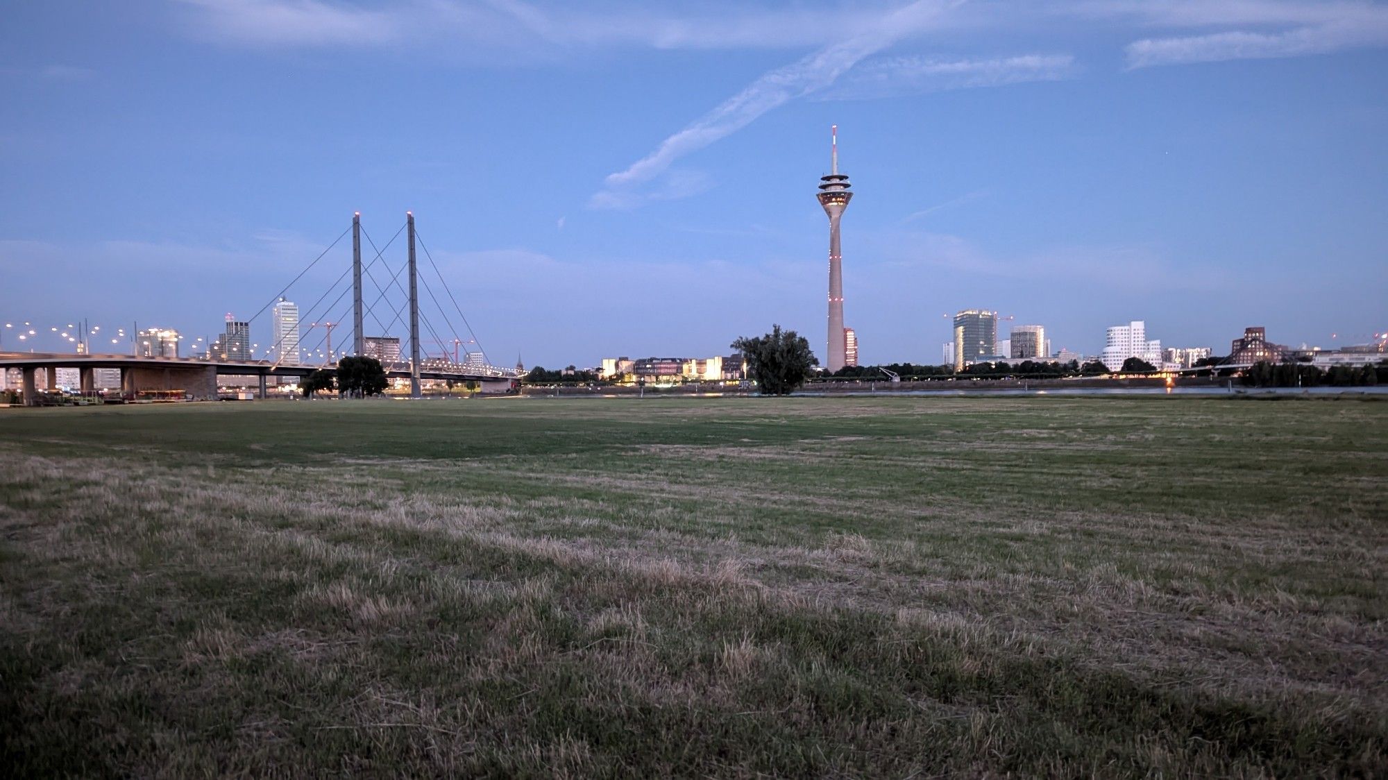 Die Skyline von Düsseldorf vom Rheinknie aus, Blick über die Rheinwiesen, links die Rheinkniebrücke. Sonnenuntergang. Himmel mit Schlierenwolken.