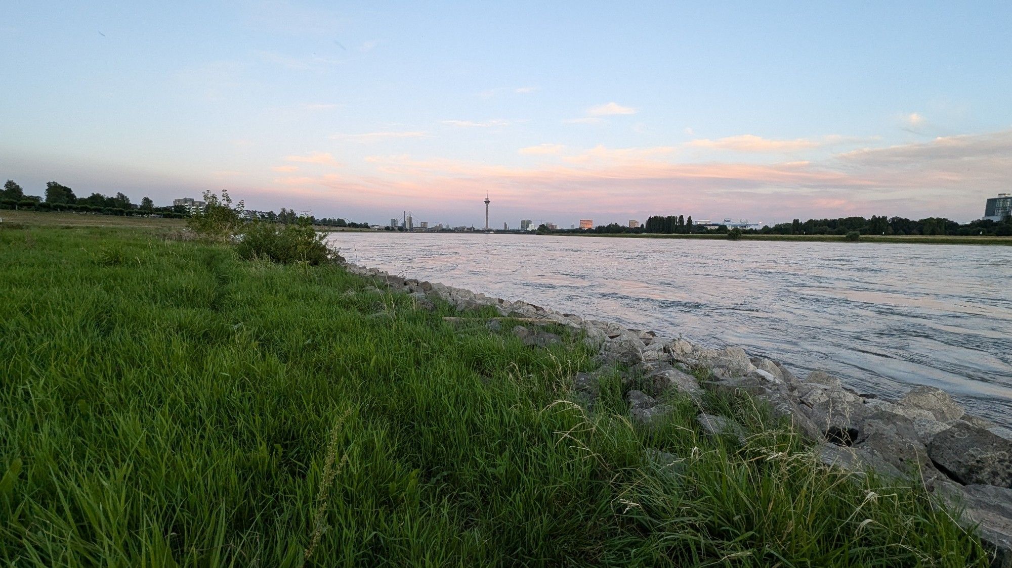 Vorne rechts bis hinten rechts und mittig der Rhein im Abendlicht, wie er quasi in die Skyline von Düsseldorf mündet. Links vorne bis hinten die Rheinwiesen und ein paar Baum-und Haussilhouetten von Düsseldorf-Oberkassel. Dahinter der Abendhimmel wie zuvor beschriebenen.