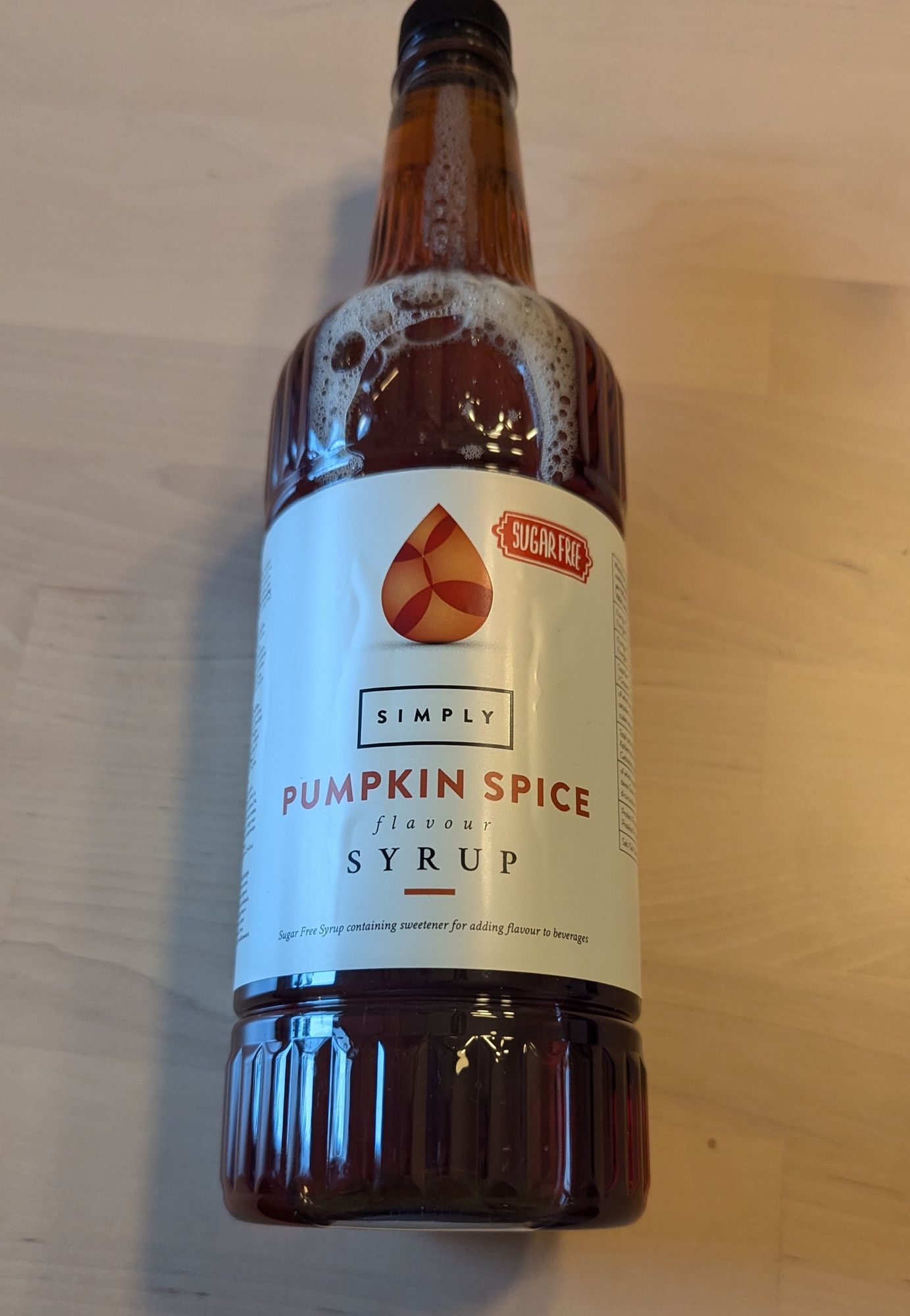 Eine Flasche Simply Pumpkin Spice Sirup, zuckerfrei, liegt auf einem hellen Holztisch. Die Flasche ist groß und braun, das Etikett weiß mit orangefarbenen Akzenten. Der Sirup ist für die Aromatisierung von Getränken gedacht.