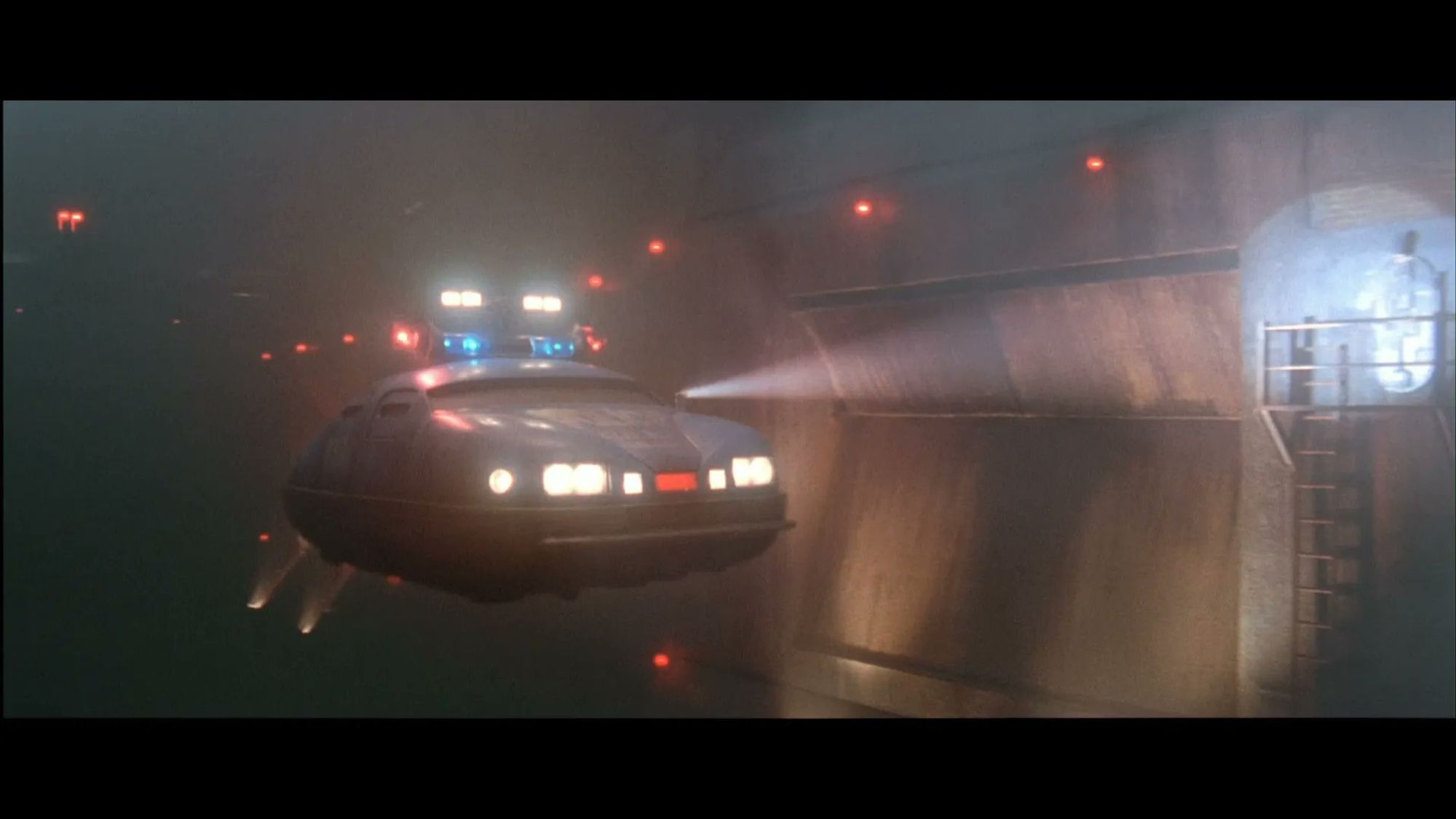 A futuristic police car, hovering above a dark, watery tunnel. The car's lights are on, illuminating the tunnel ahead. The tunnel walls are metal and damp-looking.