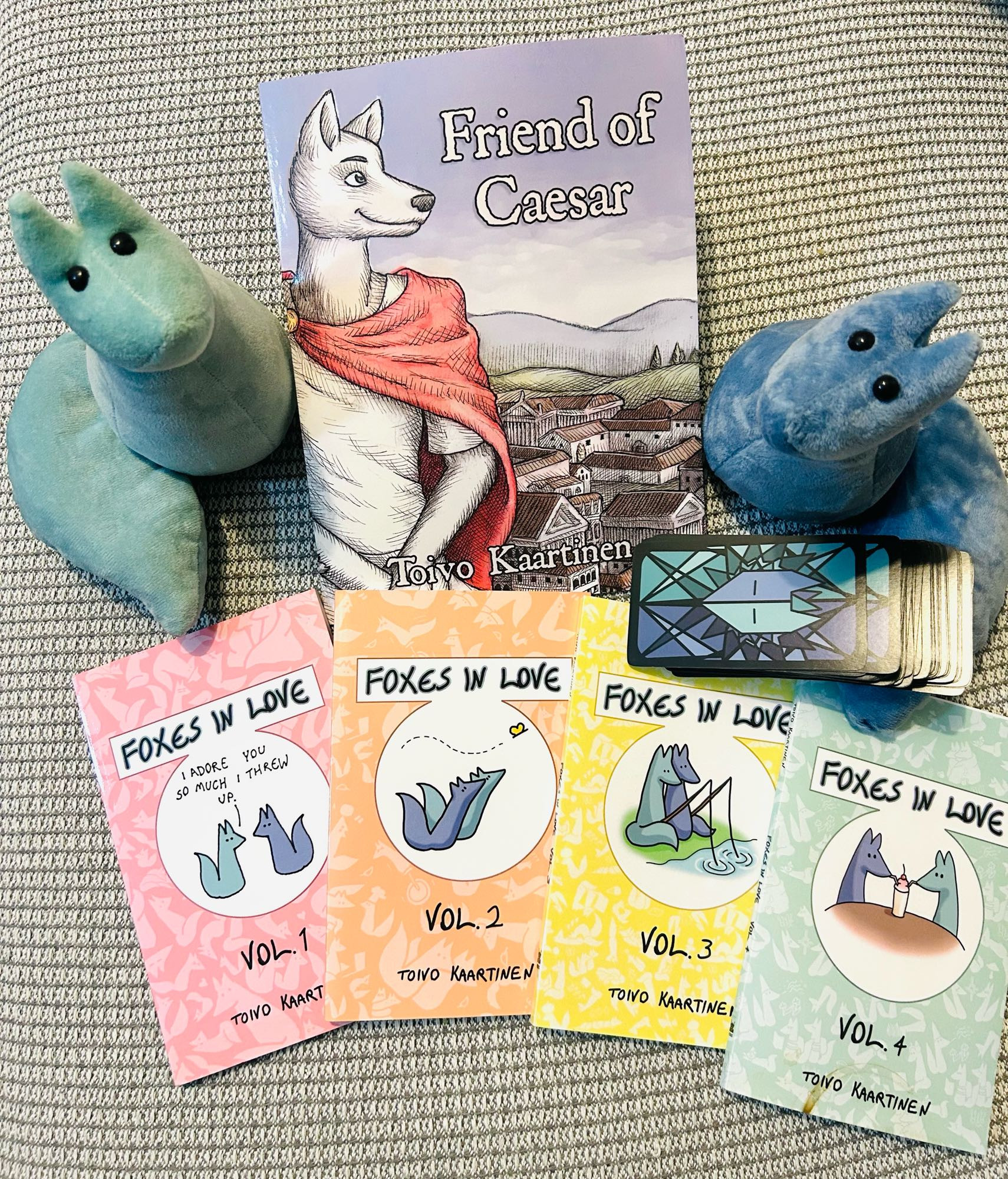 My own collection of the Foxes In Love merch produced by Fenris Publishing: 4 albums of Foxes In Love comics, the first book of Toivo Kaartinen’s unrelated series Friend of Caesar, a pack of Foxes in Love tarot deck, and Green and Blue fox plushies.