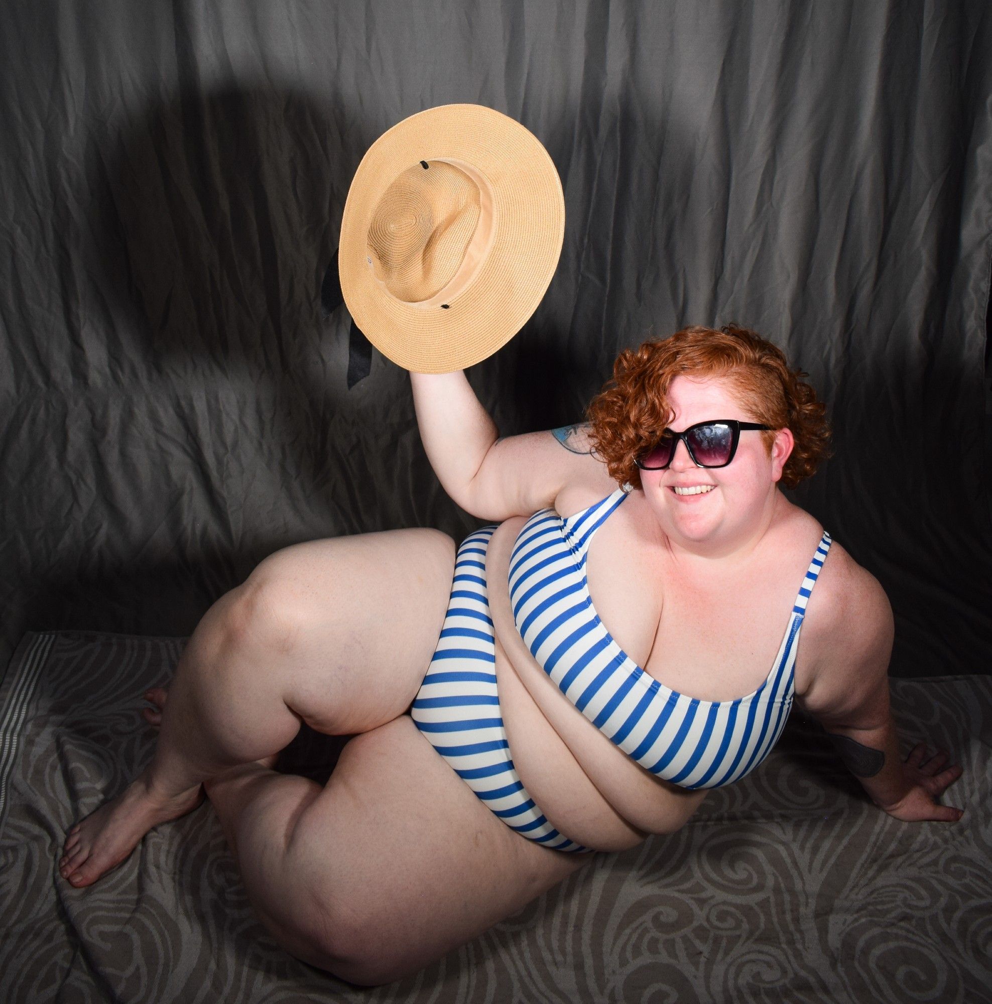 a fat woman with red curly hair in a blue and white striped two piece bathing suit, sunglasses, and a sun hat