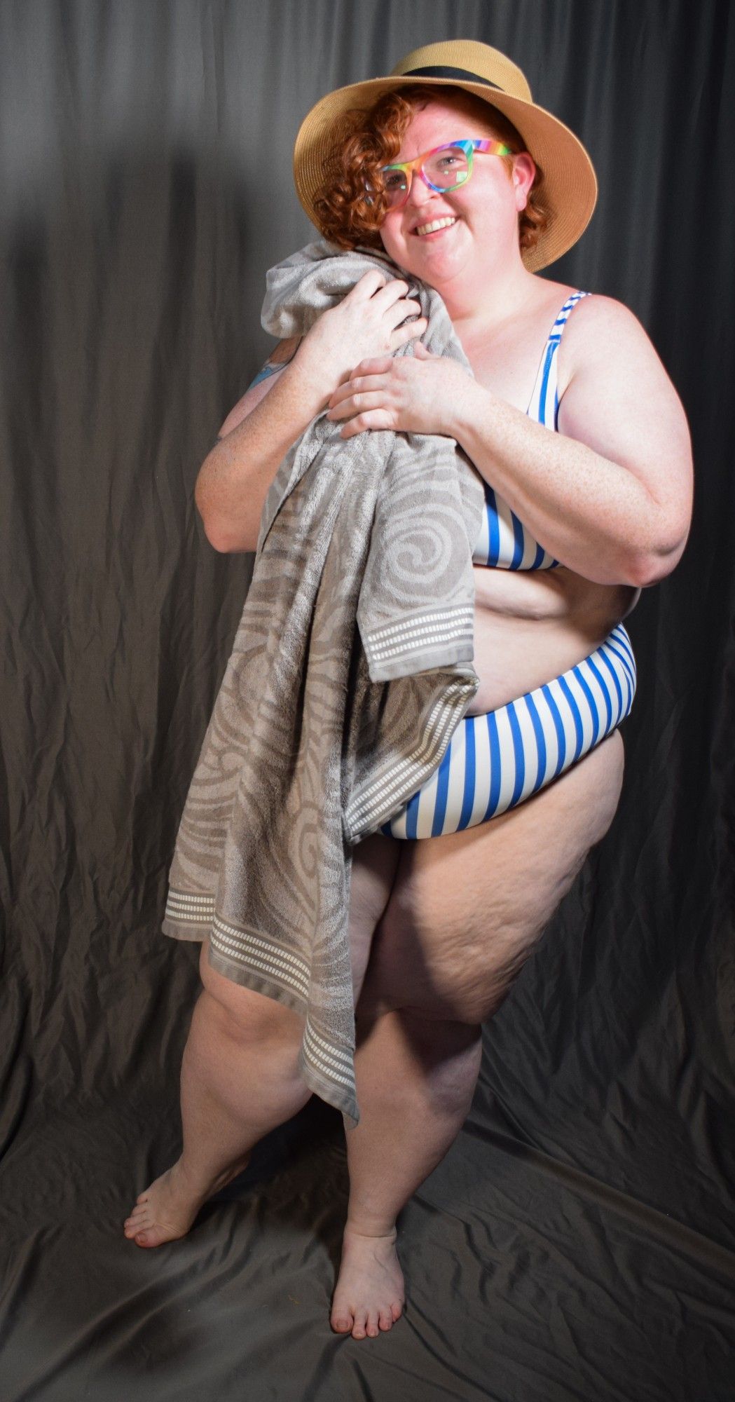 a fat woman with red curly hair in a blue and white striped two piece bathing suit, sunglasses, and a sun hat