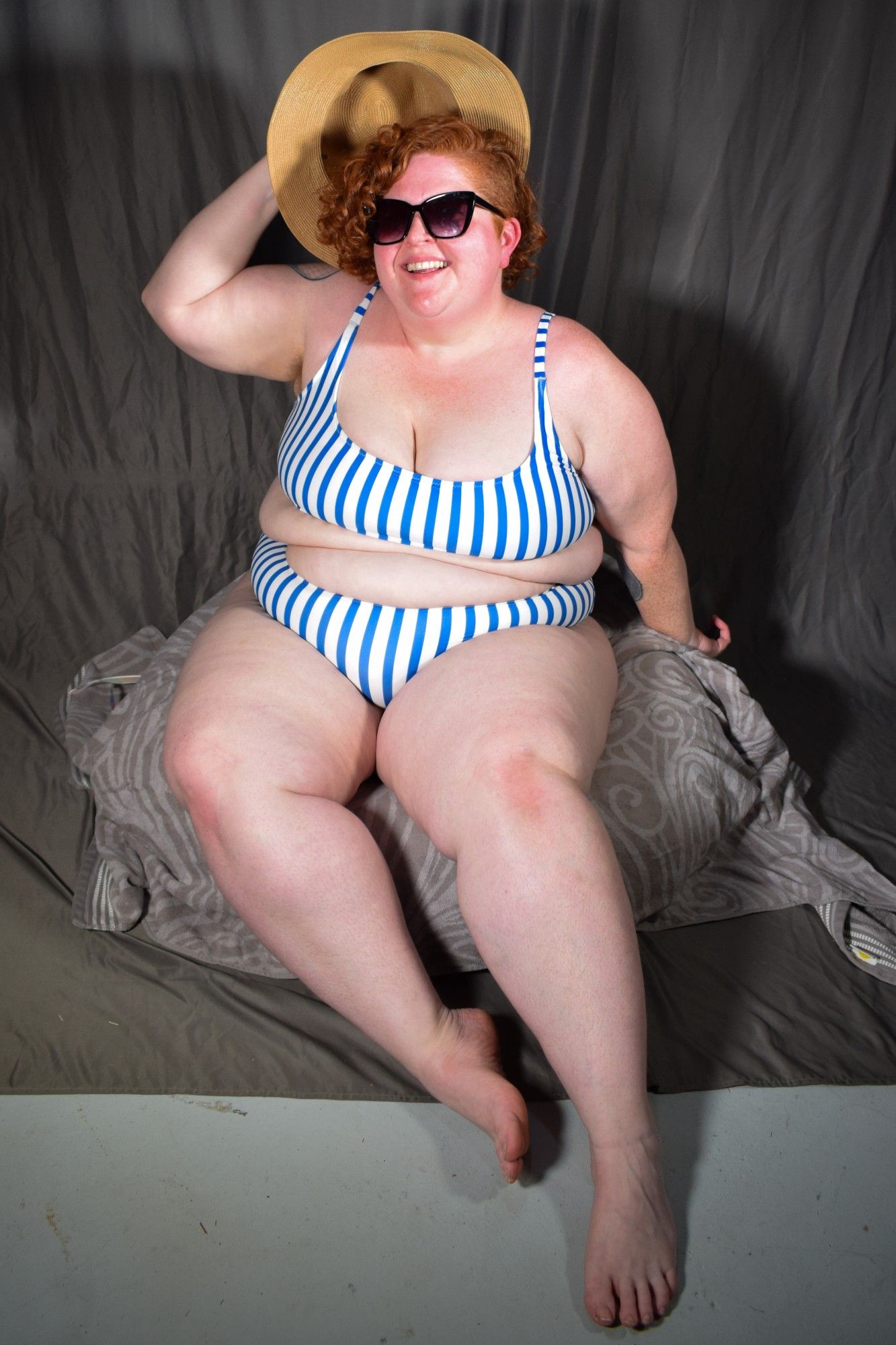 a fat woman with red curly hair in a blue and white striped two piece bathing suit, sunglasses, and a sun hat