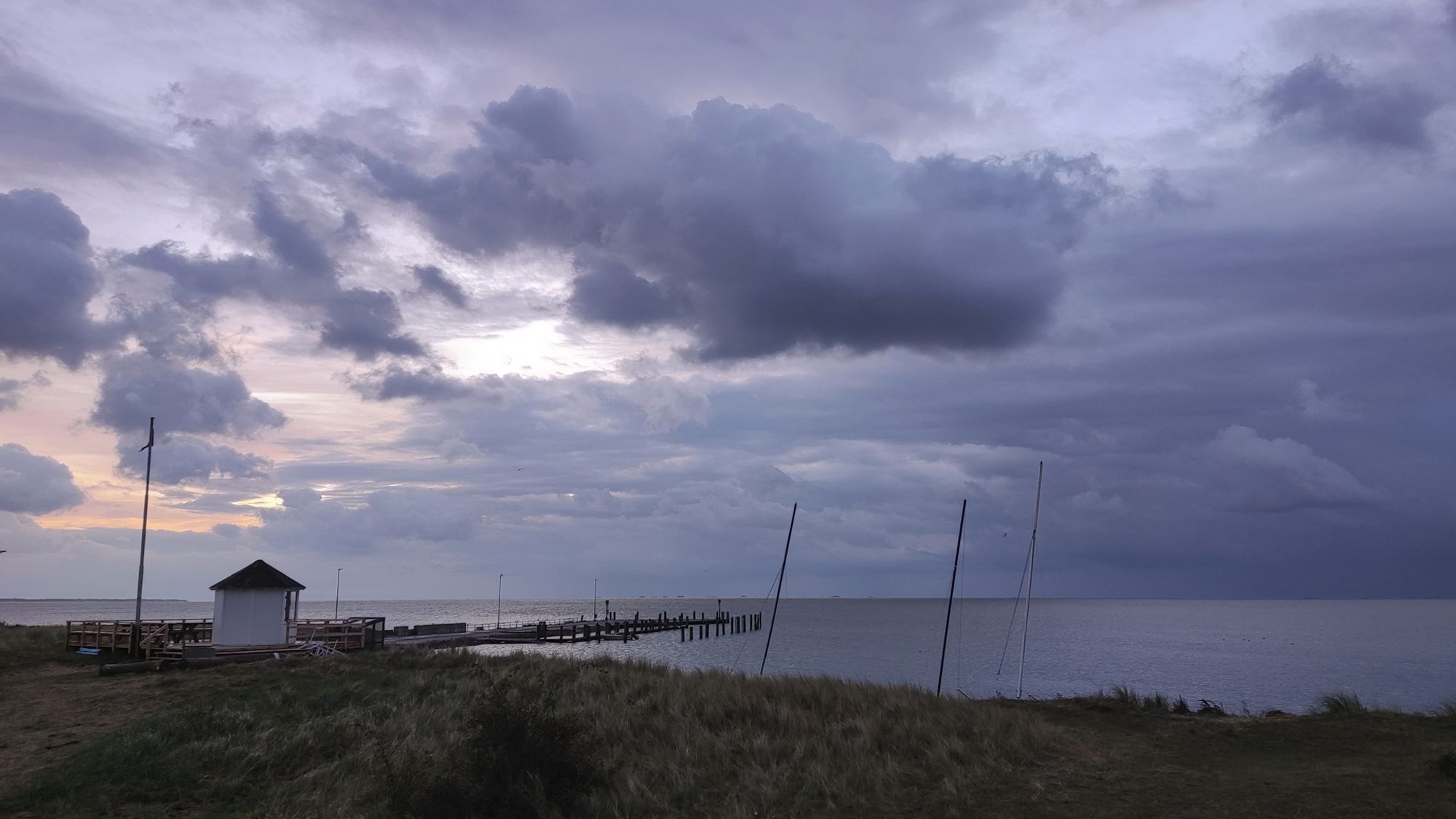 Eine Bucht am Meer. Links steht ein kleines weißes Häuschen. Man sieht Masten von Segelbooten. Der Himmel ist dunkel bewölkt.