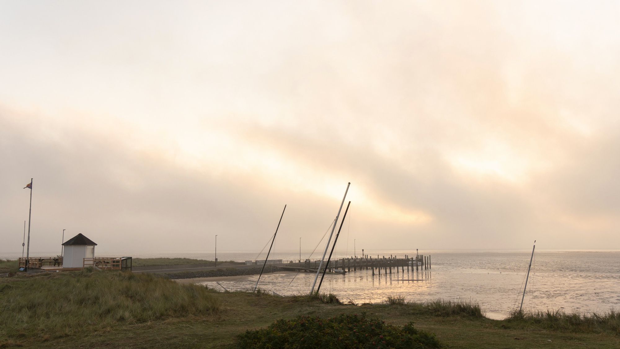 Eine Bucht am Meer. Links steht ein kleines weißes Häuschen. Man sieht Masten von Segelbooten. Der Himmel ist diffus bewölkt.