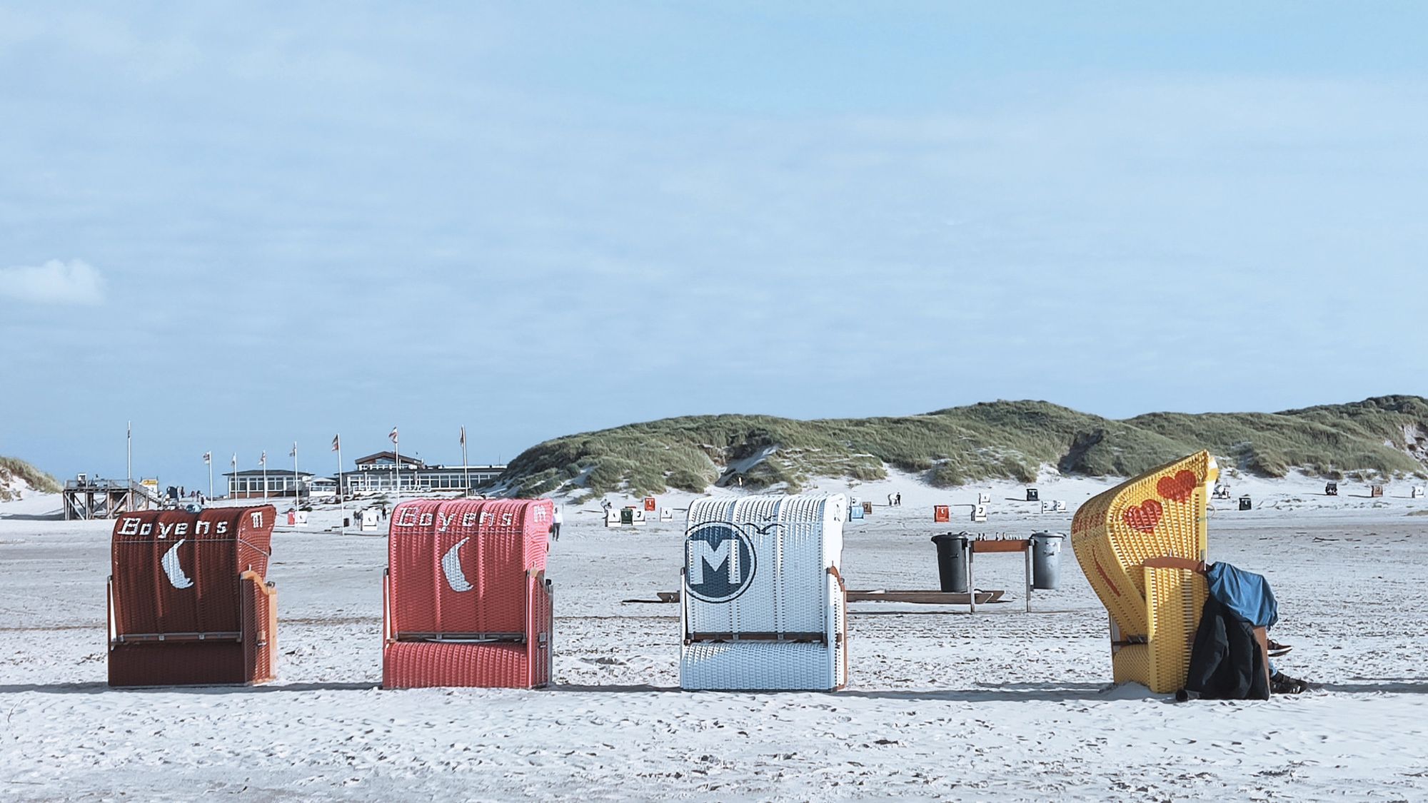 4 Strandkörbe in verschiedenen Farben stehen am Strand. 3 mit dem Rücken zur Fotografin, einer seitlich. Im Hintergrund sind Dünen.