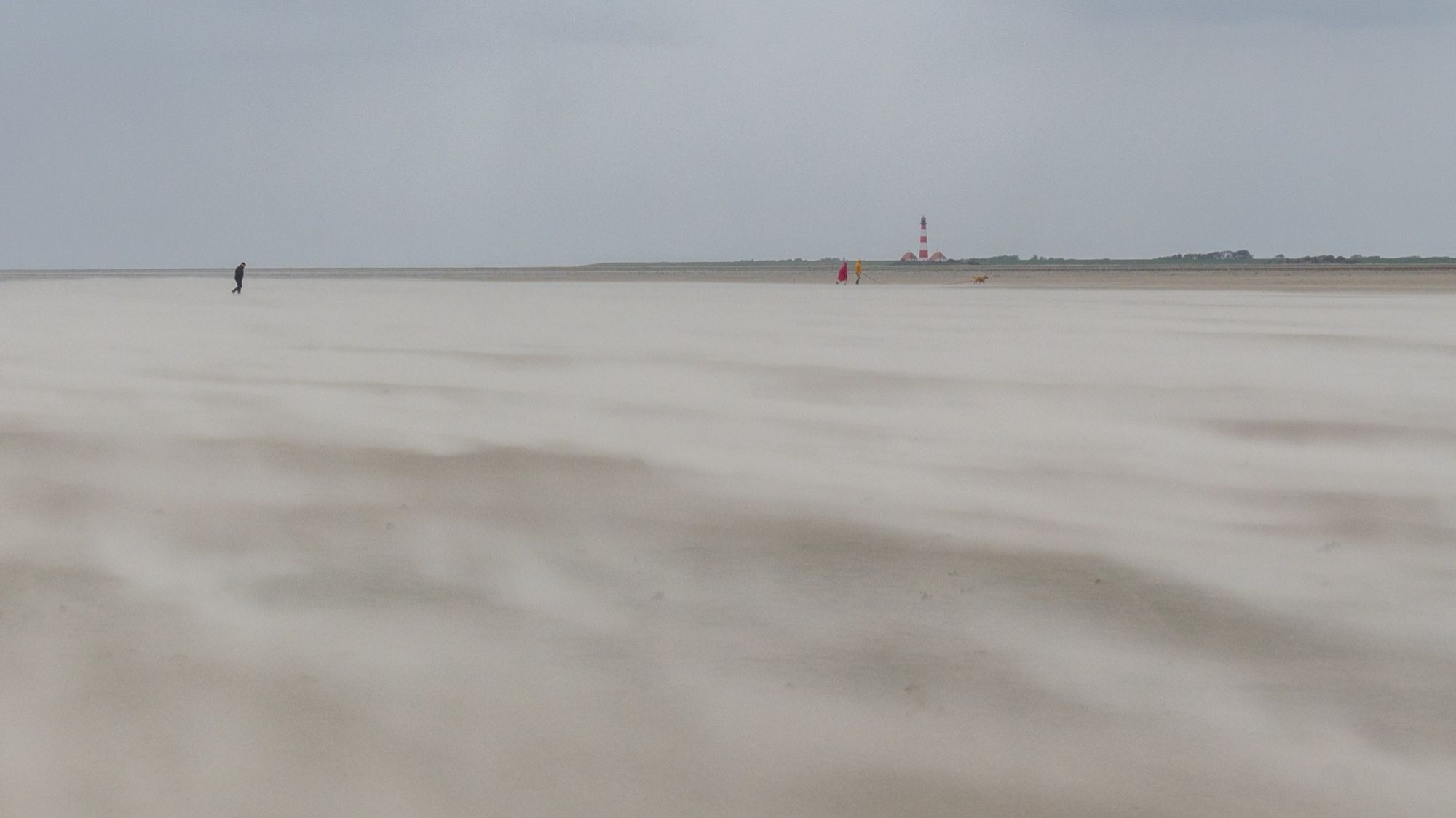 Ein großer Strand, über den der Sturm den Sand treibt. Links am Horizont ein einsamer Spaziergänger. Rechts zwei weitere Spaziergänger und ein Leuchtturm.