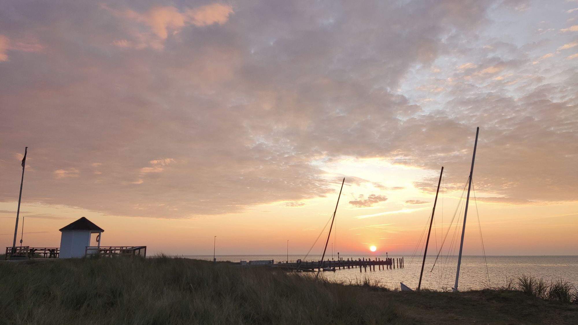 Eine Bucht am Meer. Links steht ein kleines weißes Häuschen. Man sieht Masten von Segelbooten. Die Sonne geht auf, der Himmel ist teilweise bewölkt.