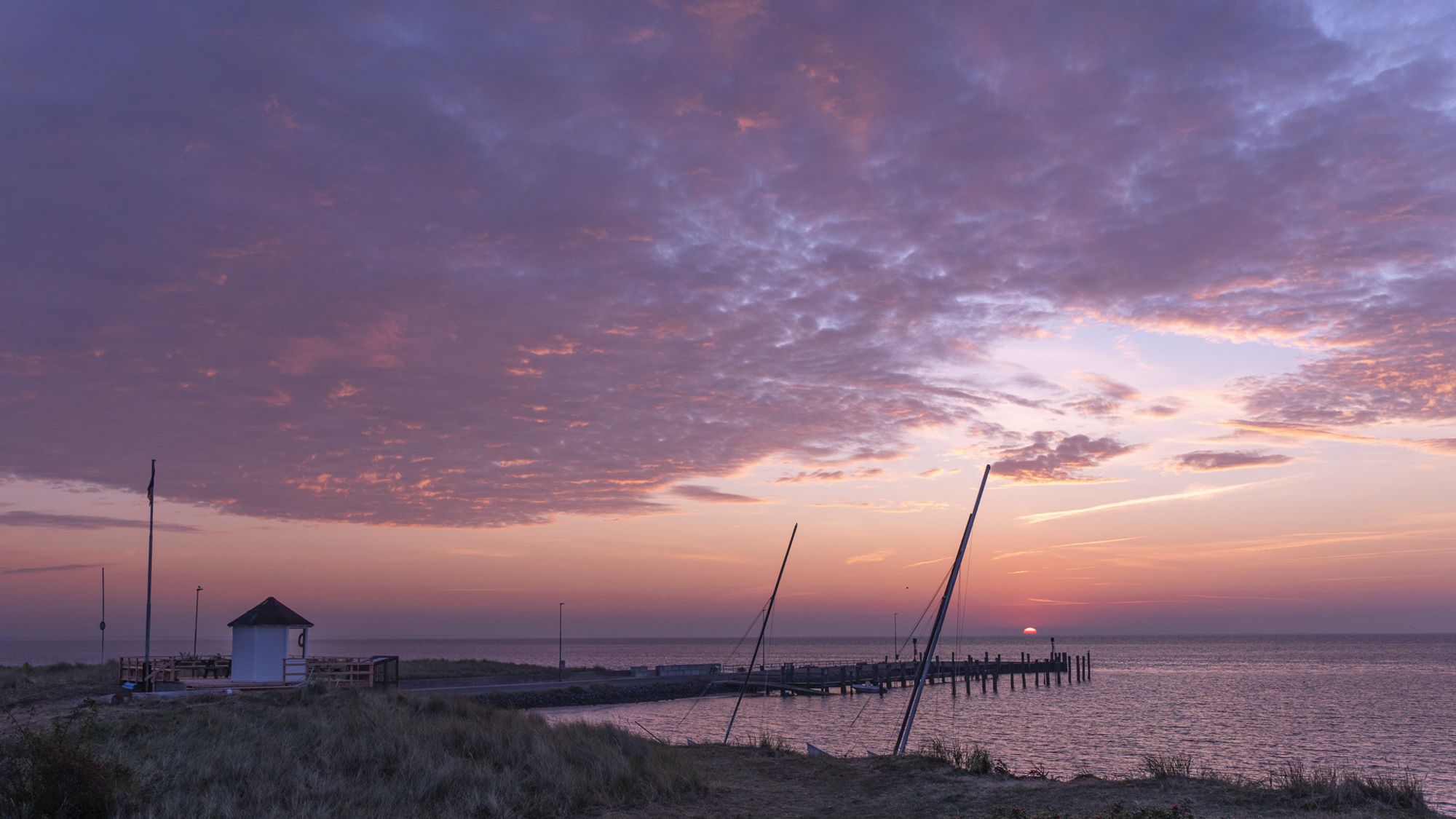 Eine Bucht am Meer. Links steht ein kleines weißes Häuschen. Man sieht Masten von Segelbooten. Die Sonne geht auf, der Himmel ist teilweise bewölkt.