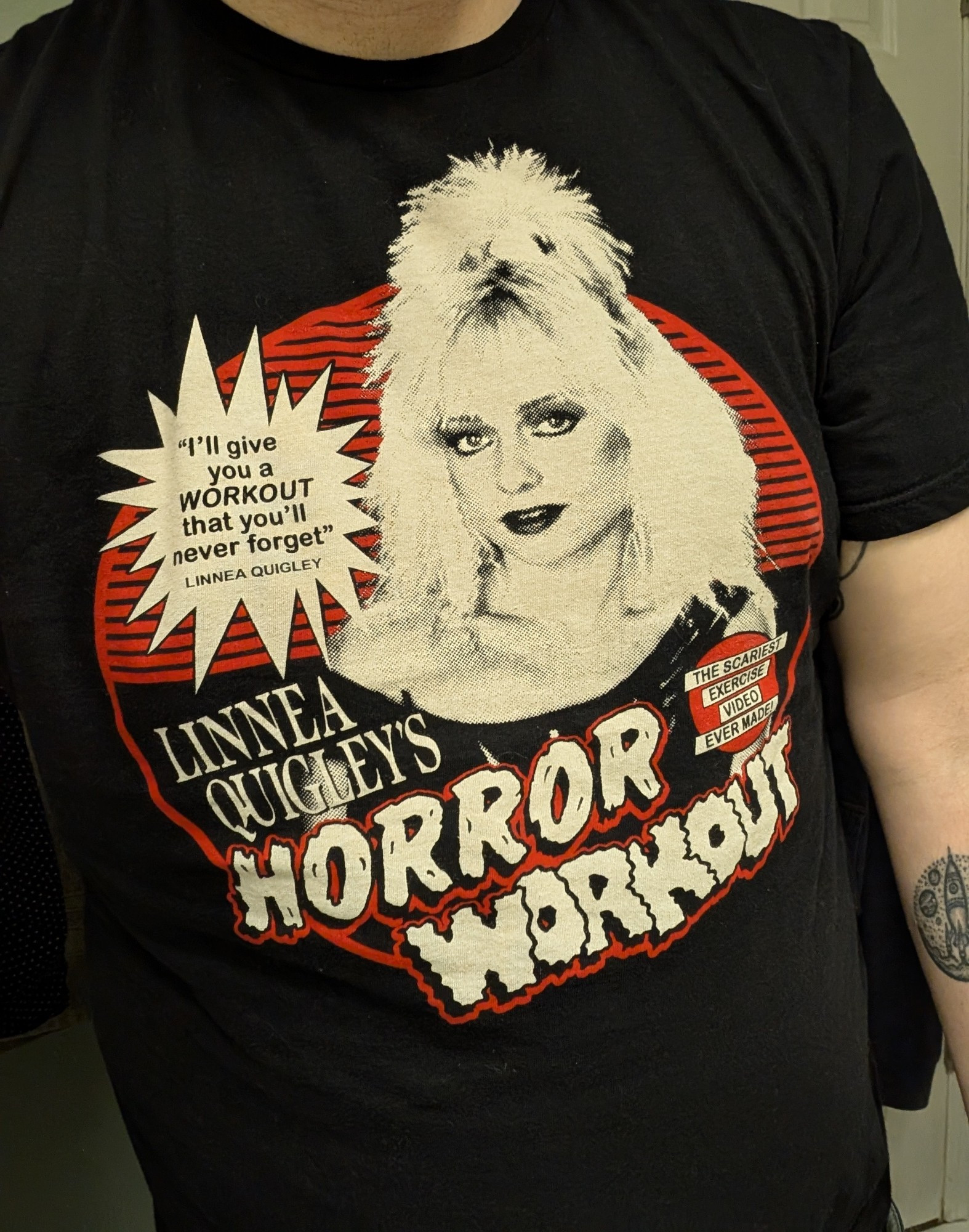 A black T-shirt with an image of actress Linnea Quigley that says "Linnea Quigley's Horror Workout". A starburst has text inside it that says "I'll give you a WORKOUT that you'll never forget!" There is also a red circle with text inside that says "The scariest exercise video ever made!"