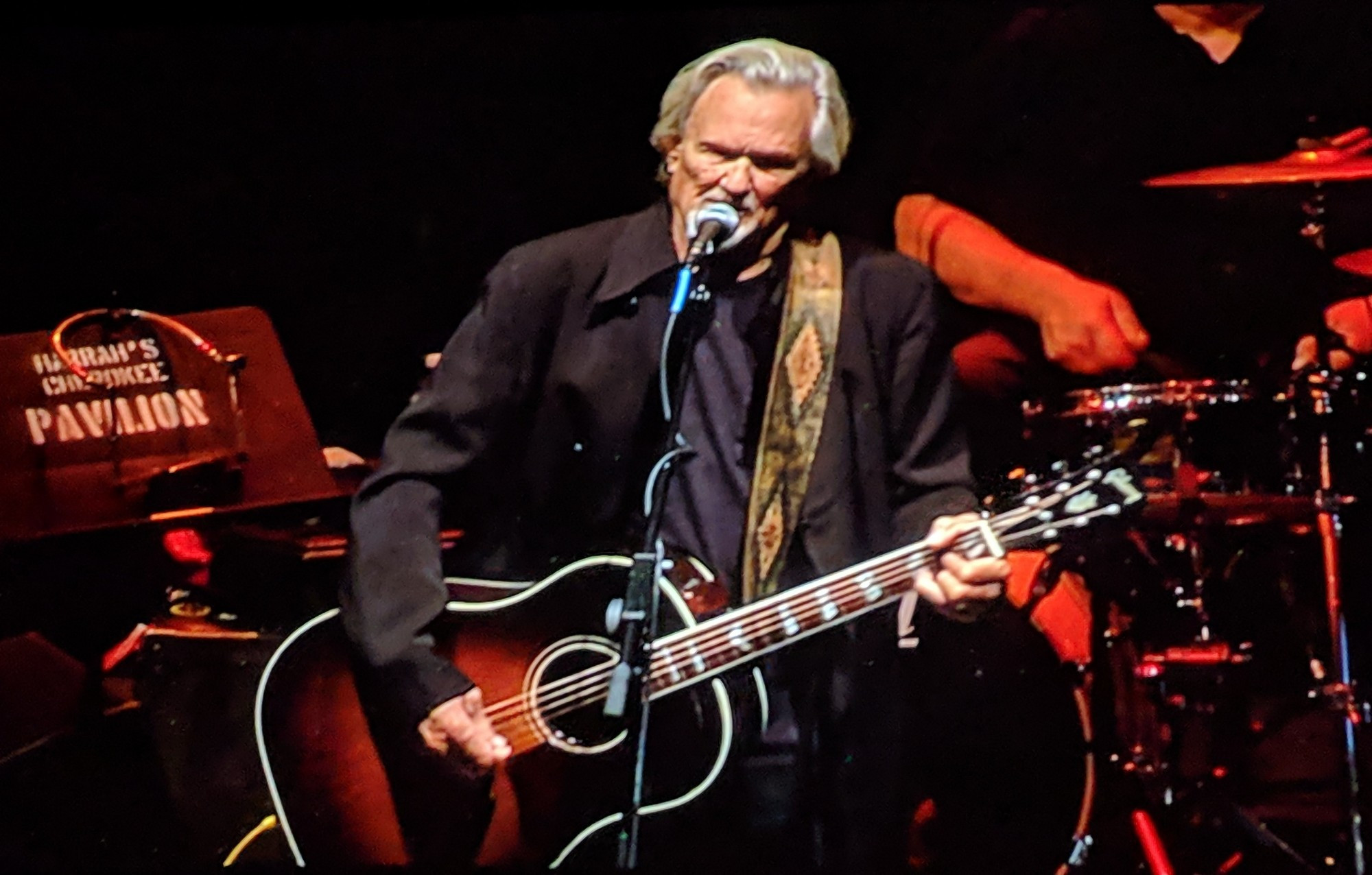 Kris Kristofferson performing at Harrah's Casino in Cherokee, NC in 2018. Image shows Kristofferson standing at a mic so ging and playing guitar.