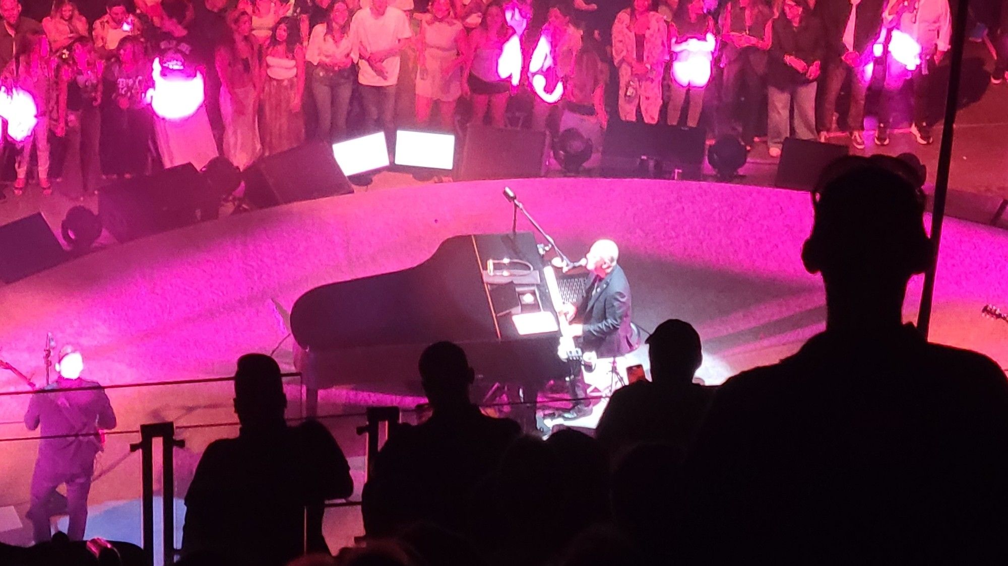 Billy Joel, on stage, seated at his piano bathed in pink light.