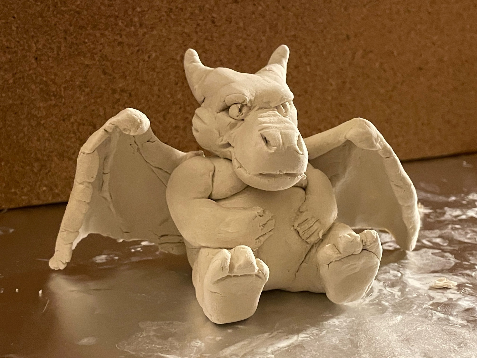 An image of a small clay humanoid dragon with teddy bear porportions, sitting peacefully with his hands on his round belly, on a messy sheet of tinfoil with a corkboard backdrop.