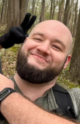 An image of a bald man with a beard wearing a green shirt and backpack. 