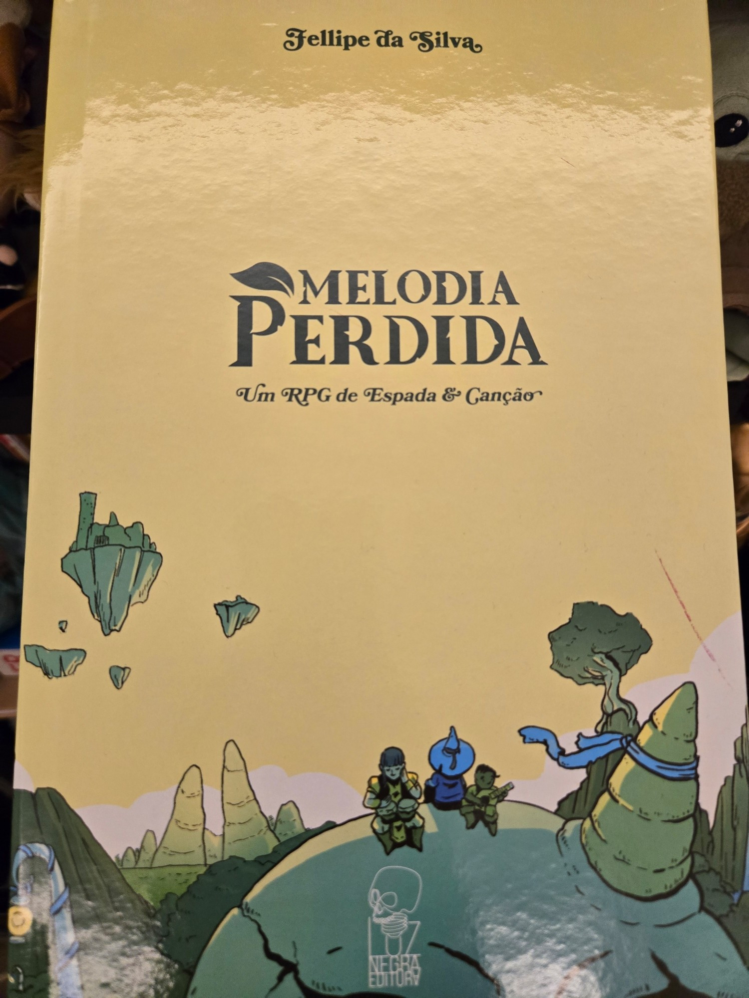 A imagem mostra a capa do livro Melodia Perdida: Um RPG de Espada & Canção, escrito por Fellipe da Silva. O título está em destaque no centro da capa, com um design que remete à fantasia clássica. A arte apresenta um cenário com tons pastéis e estilizados, mostrando um grupo de aventureiros sentado em um ambiente fantástico, com formações rochosas e ilhas flutuantes ao fundo. Os personagens possuem um estilo de ilustração cartunesco e estão em um momento de descanso ou contemplação, sugerindo um tom de aventura com momentos de introspecção e música. No canto inferior direito, há o logotipo da "Luz Negra Editora". A capa transmite um clima de fantasia e exploração, com foco em narrativas envolventes que misturam combate e música.