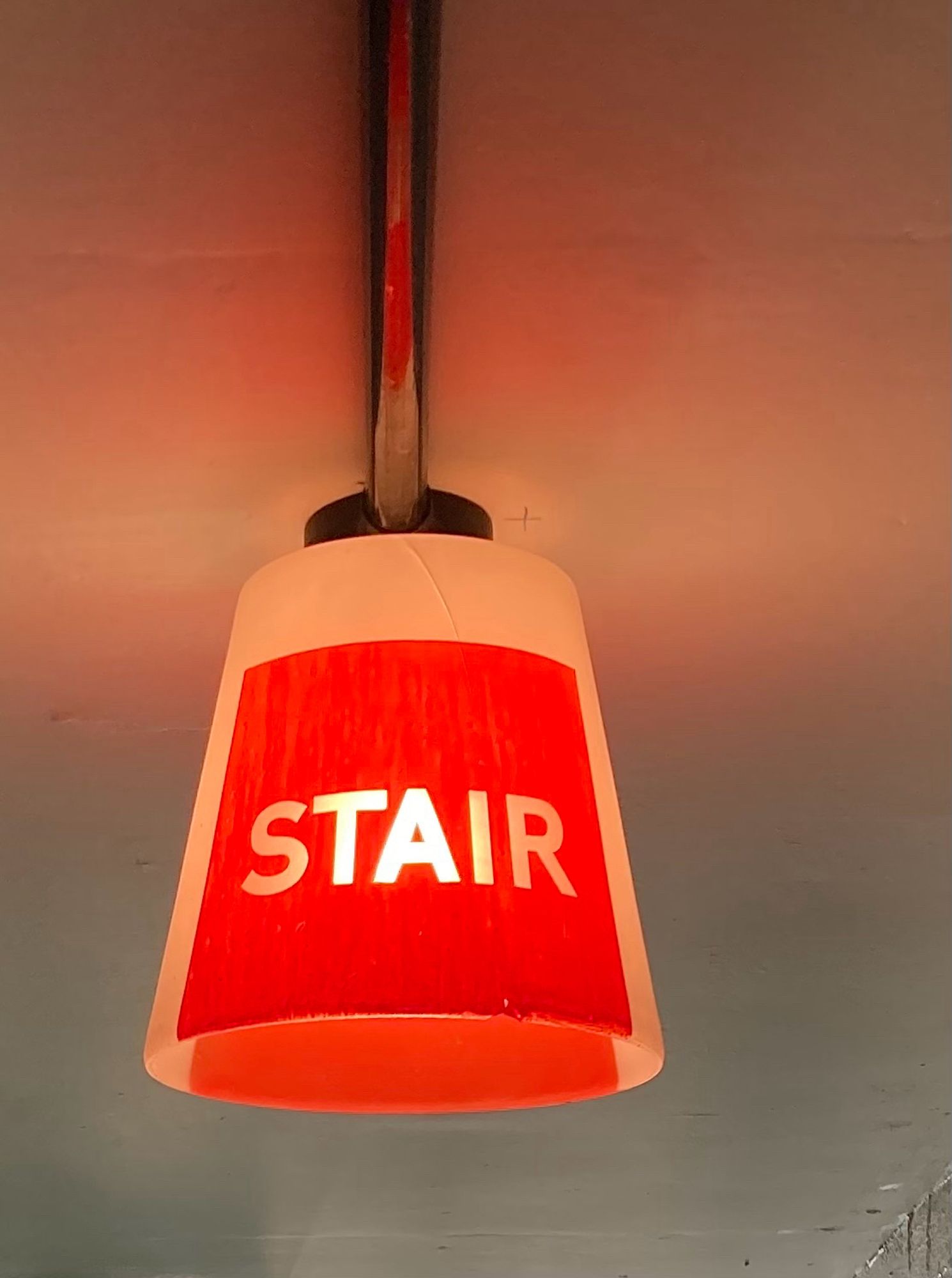 A 1930s glass lampshade hanging from a ceiling, with the word ‘Stair’ illuminated through a patch of red paint.