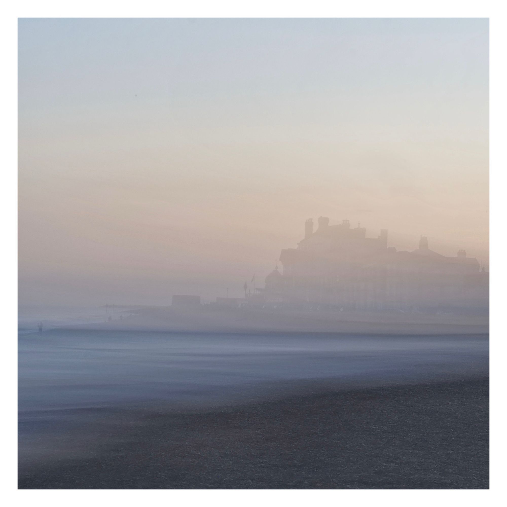 ICM photograph of soft pink evening light, the distant impression of buildings overlooking the grey blue beach and sea.