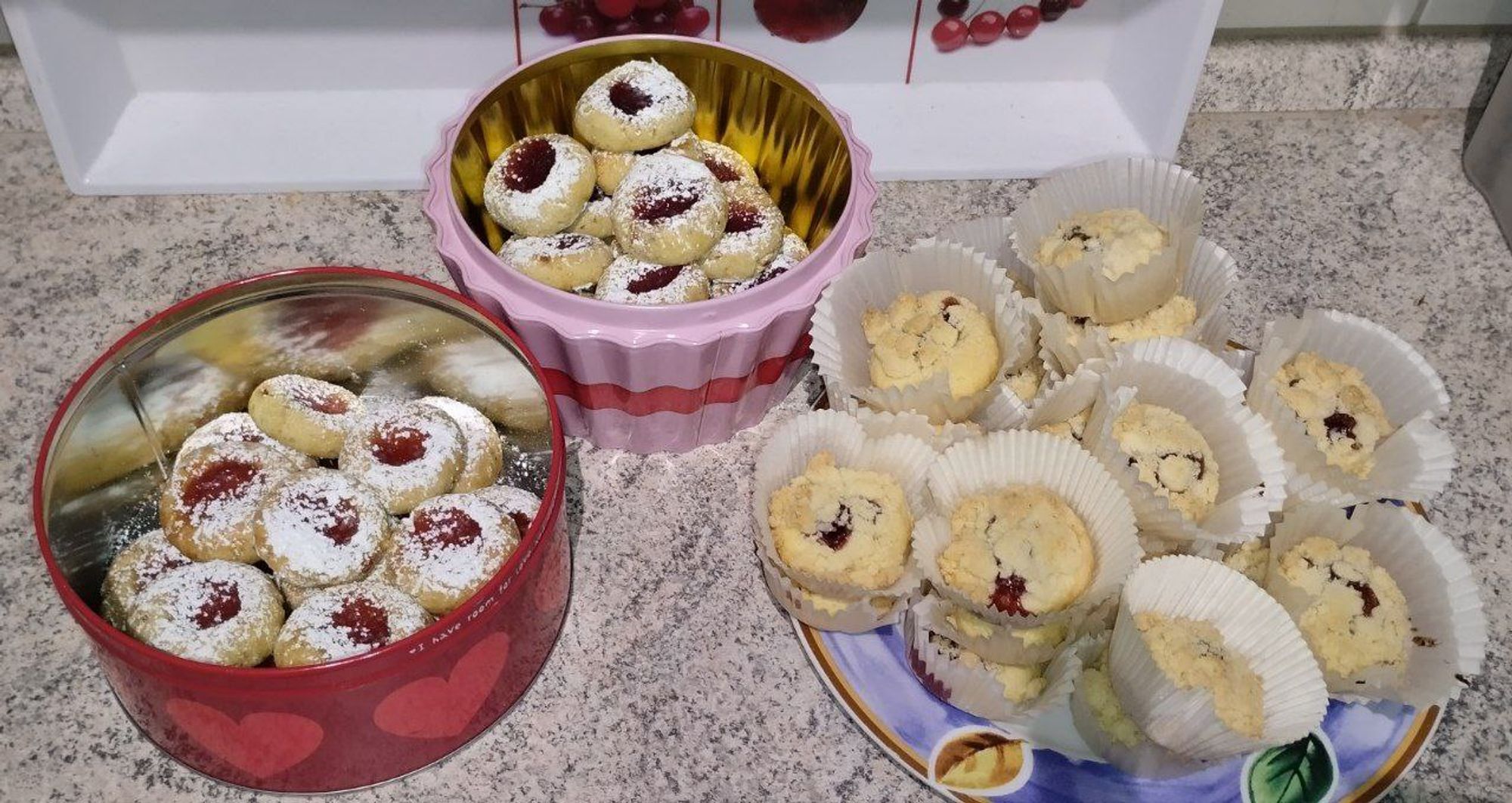 Auf einem Küchentisch stehen eine rote und eine rosa Dose je mit Husarenkrapfen, die mit roten Marmeladen gefüllt und Puderzucker bestreut sind. Daneben stapeln auf einem großen Teller mit blauem Rand viele Muffinförmchen, in denen hoch gebackene Butterkekse mit Marmelade und Streuseln sind.