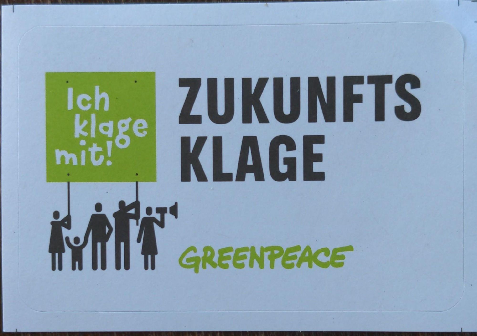 Aufkleber mit einer Gruppe von Menschen, die ein Schild hochhält mit 'Ich klage mit!' und dem Schriftzug Zukunftsklage.