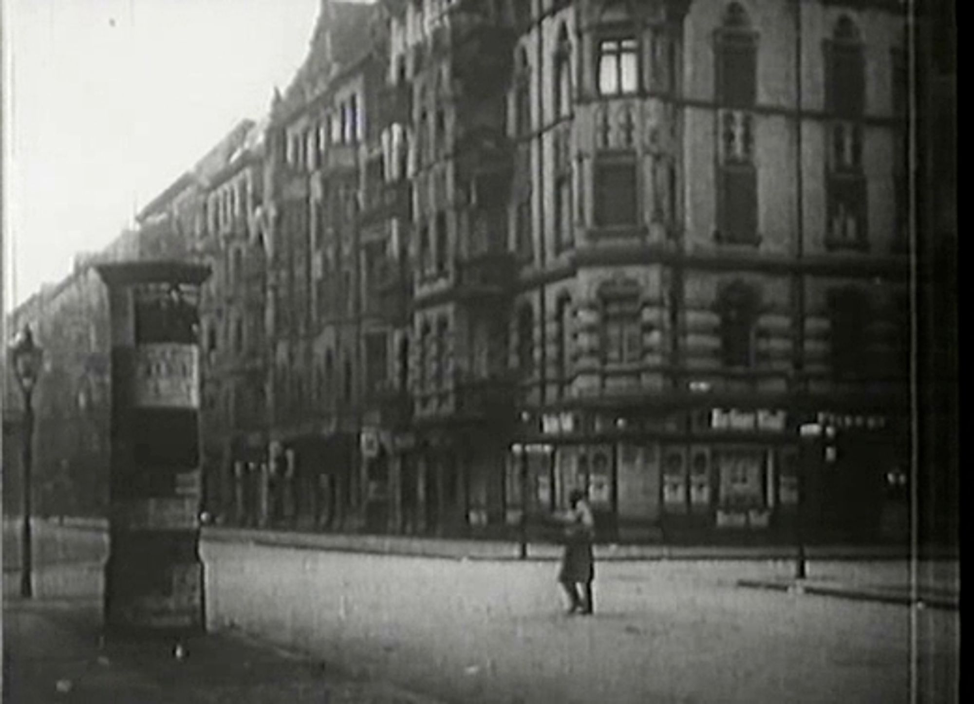 Erwachens-Szene des Films zeigt eine leere Straße, an der links eine Litfassäule steht und mittig ein einzelner Mensch über die Straße geht. Dahinter viergeschossige Häuser, rechts eine Berliner Kindl Kneipe. Nachweis von wiki commons folgt.