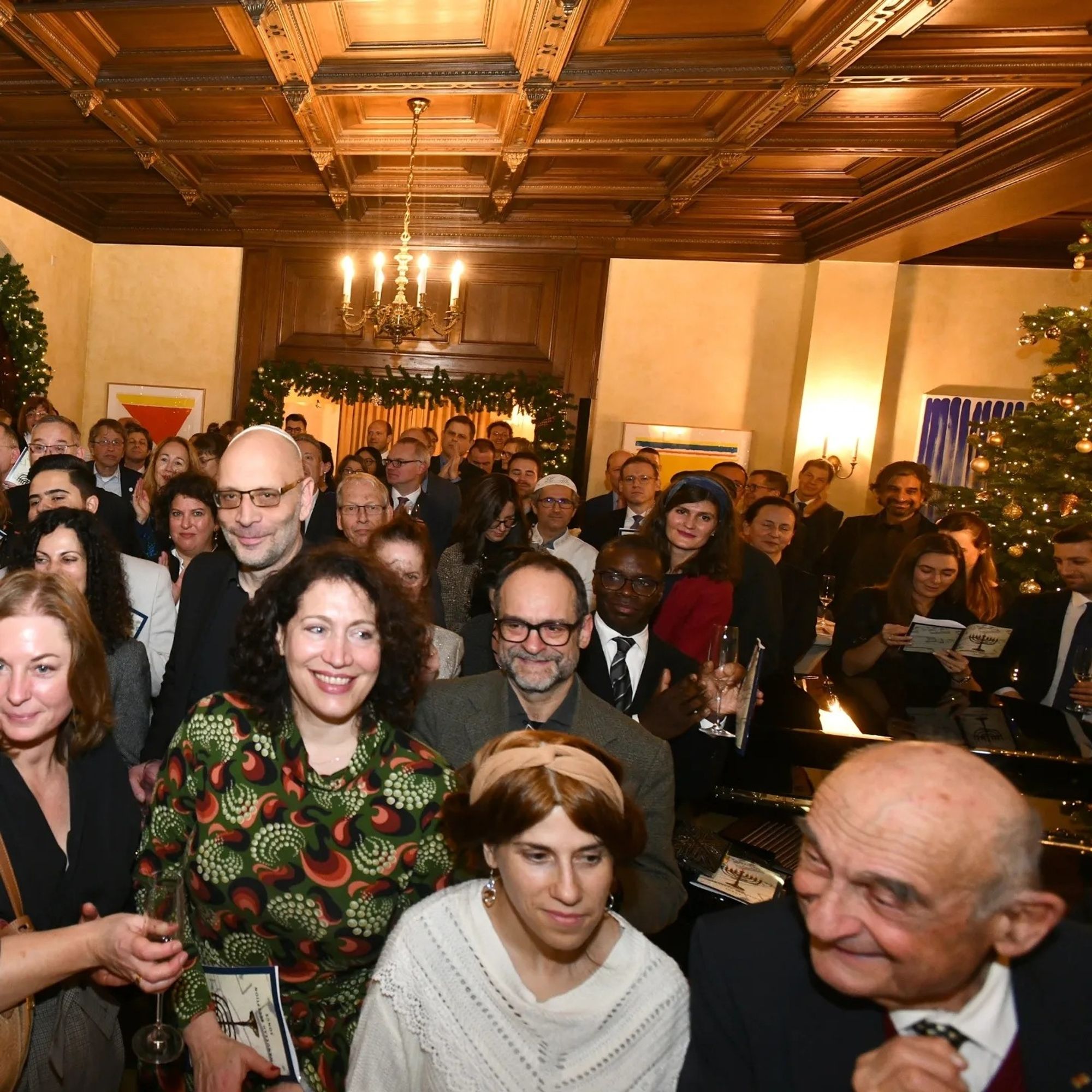 Ein holzvertäfelter Saal voller Gäste, die der Musik lauschen. Rechts ein Flügel, dahinter ein geschmückter Tannenbaum.