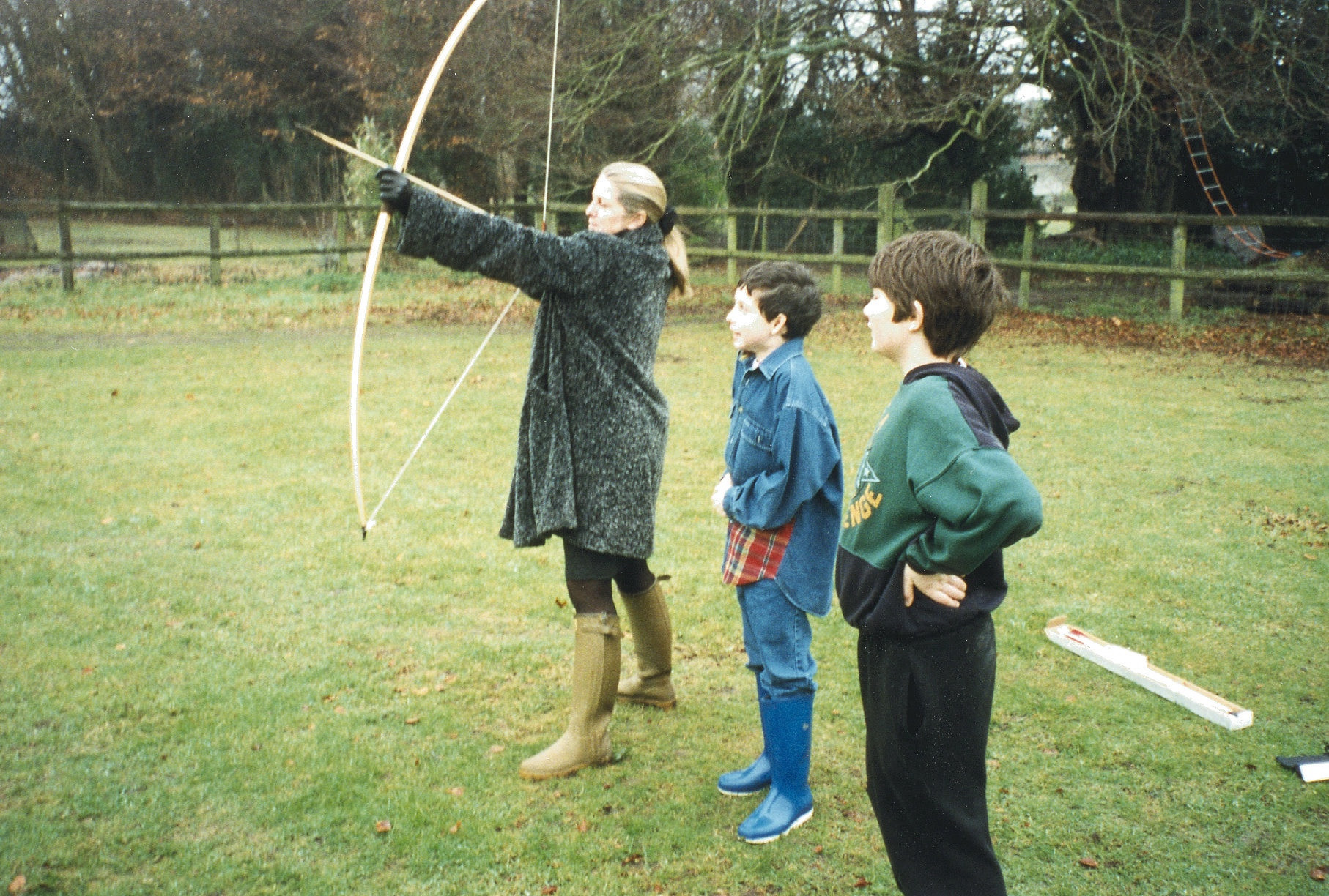 Mummy shooting arrows from a longbow watched by two small boys. 