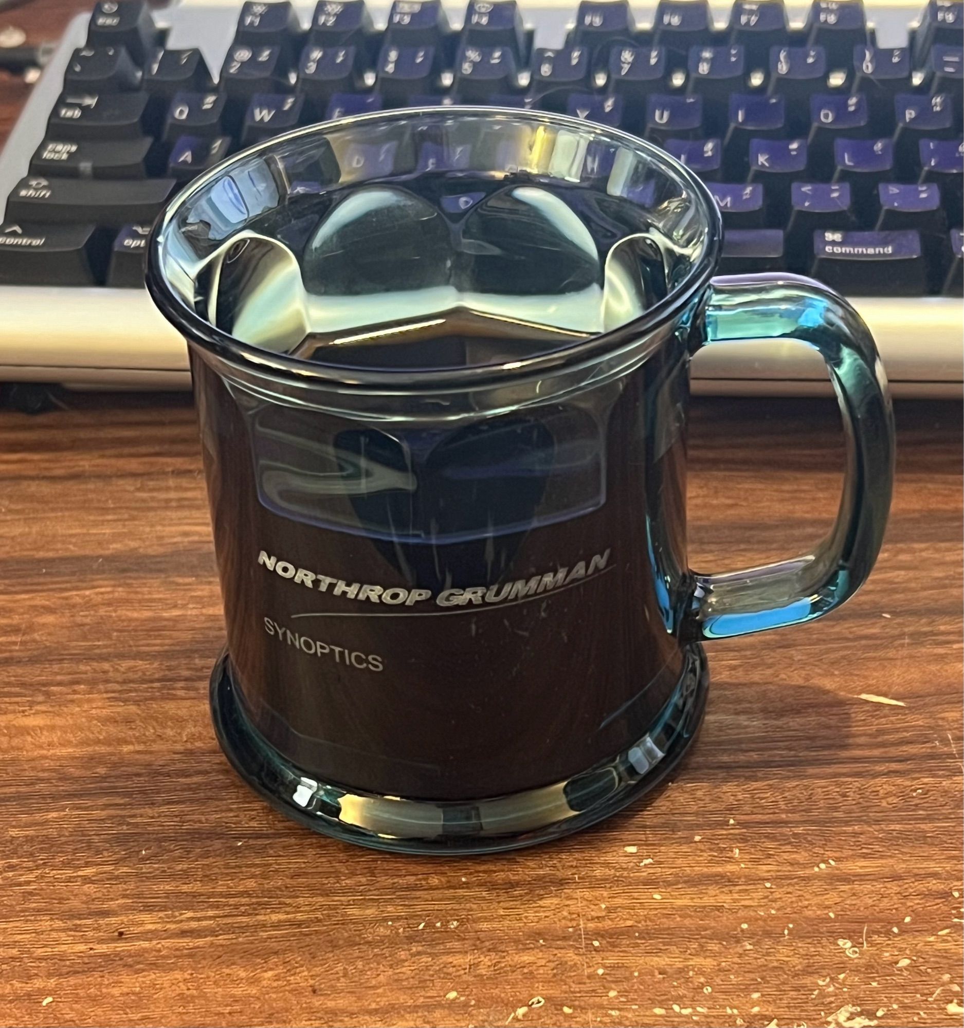 Photo of a mug of mostly full coffee on my desk. The desk is a fake wood grain and you can see my keyboard behind.

The mug is a clear blue glass with faceted inner sides and looks like a quality mug. On the front in fading silver lettering says “Northrop Grumman Synoptics”