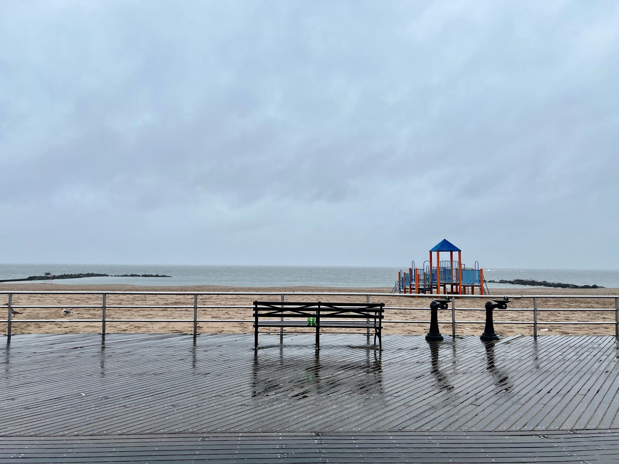 Ein Strand bei Regen. in der Mitte eine Parkbank, rechts dahinter ein Klettergerüst