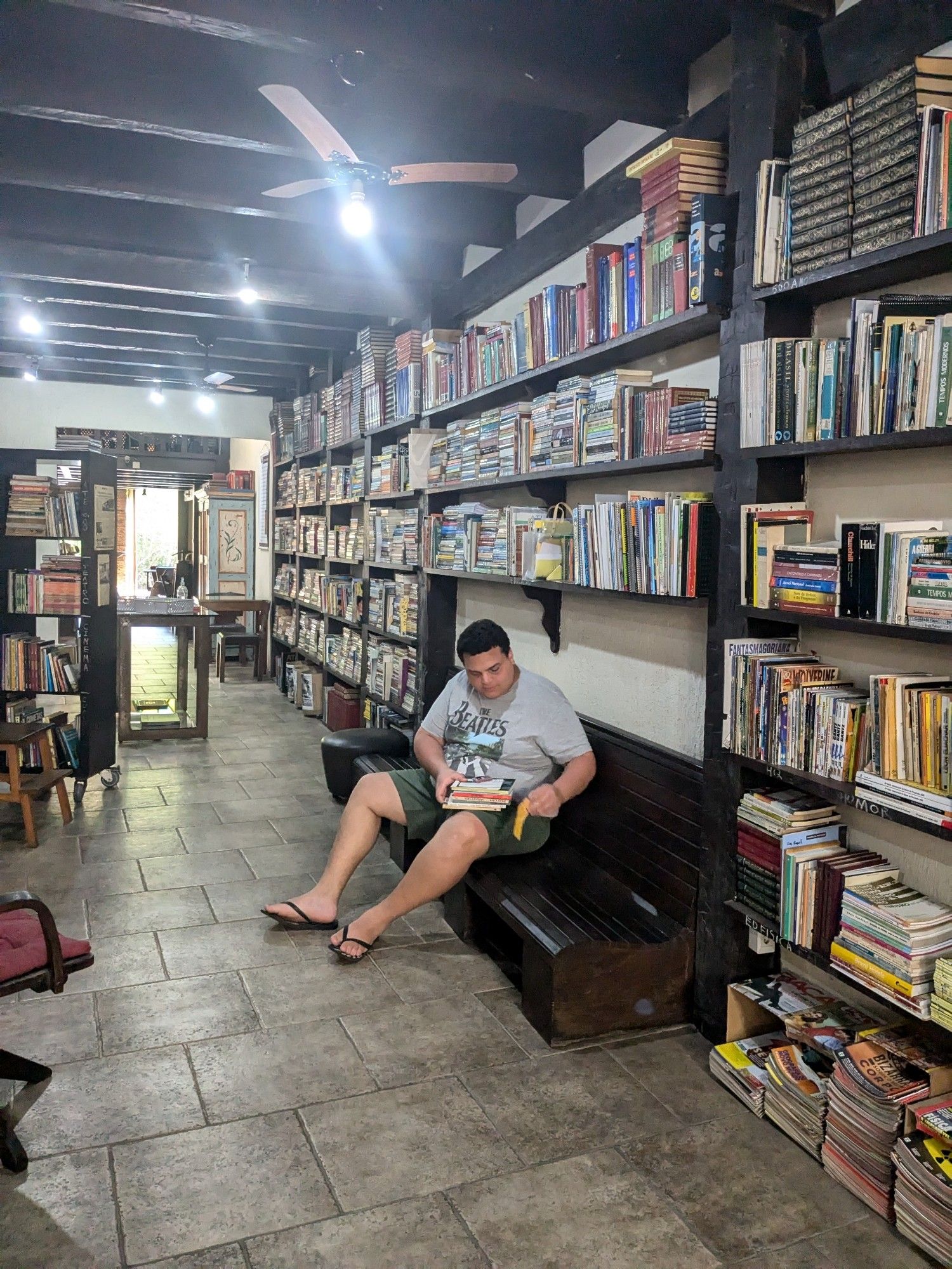 foto do nicoletti dentro do sebo "o livreiro", sentado em um banco segurando uns livros que eu escolhi. a parte interior do sebo é repleta de estantes e possui esse banco grande de madeira escura entre elas, um espaço para que os consumidores possam ler e relaxar um pouco. as paredes são brancas e as estantes, do mesmo material do que o banco (madeira escura)